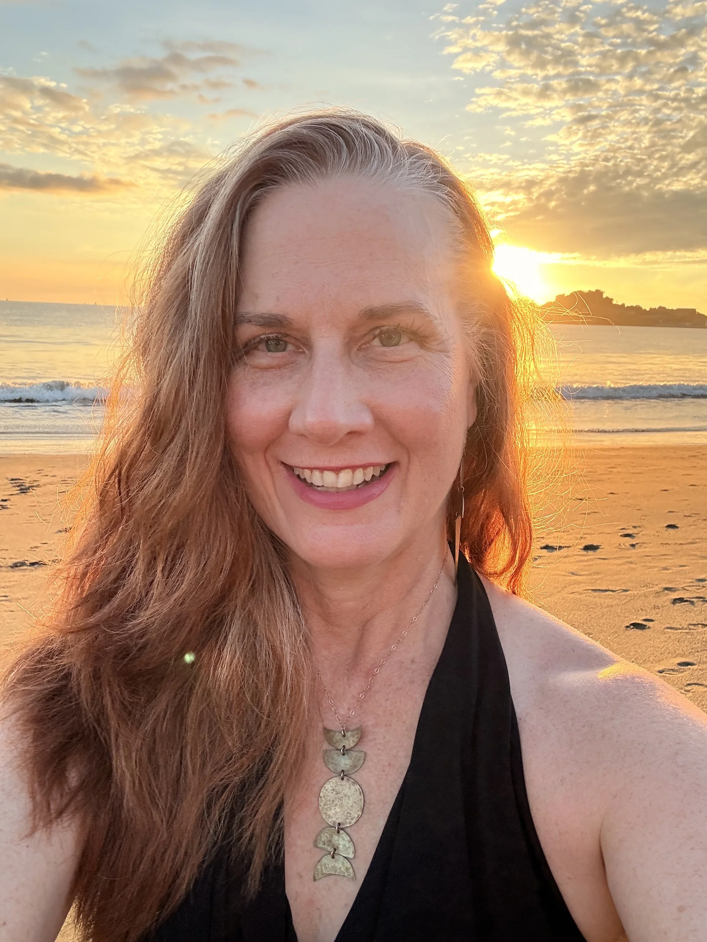 A woman with long red hair smiling at the camera on a beach during sunset. The sky is partly cloudy with orange and yellow hues, and the ocean and coastline are visible in the background.