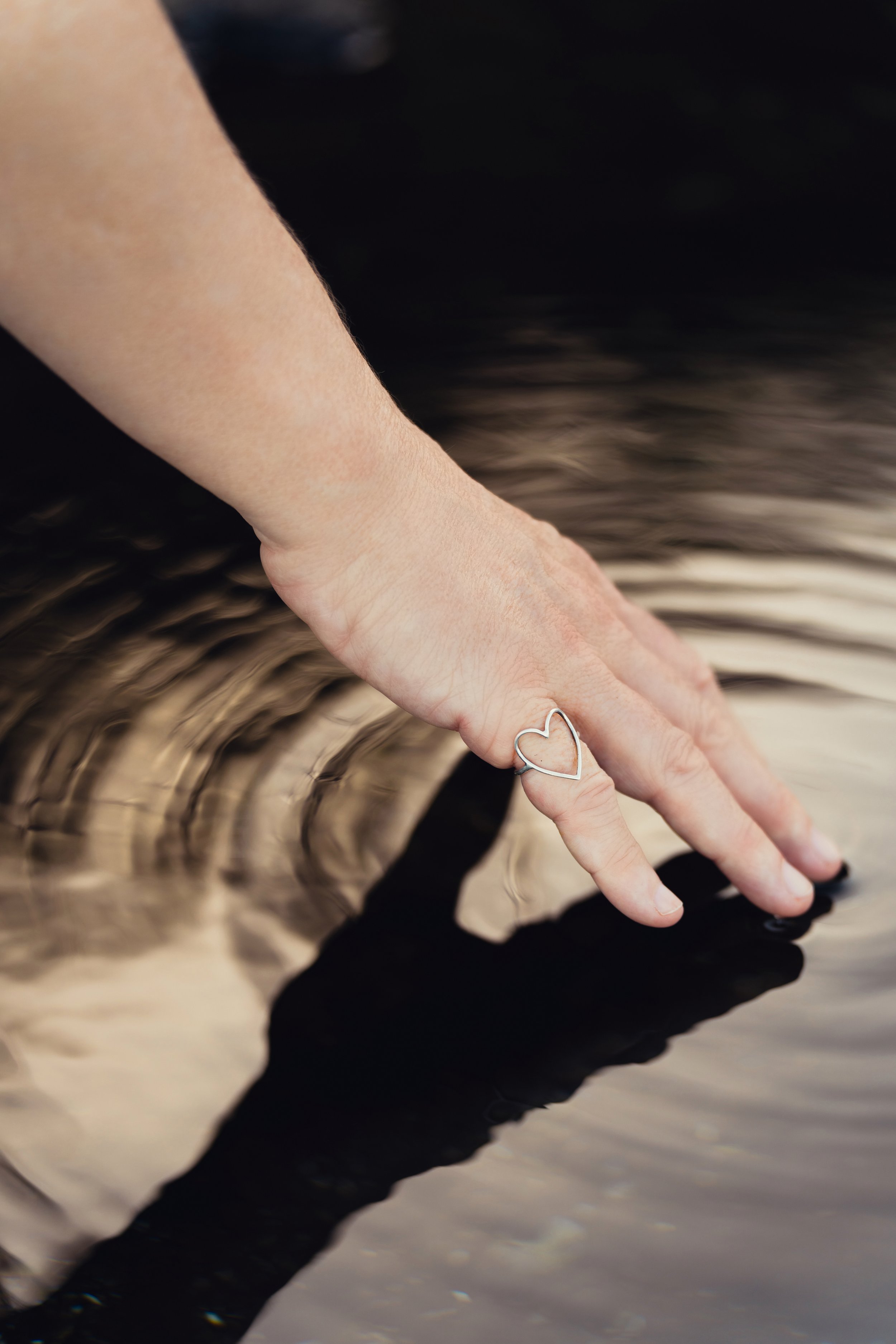 A woman's hand with a silver heart-shaped ring on the finger, touching the surface of the water in a tidal pool, making a ripple of concentric circles on the surface of the water with a blurred background.