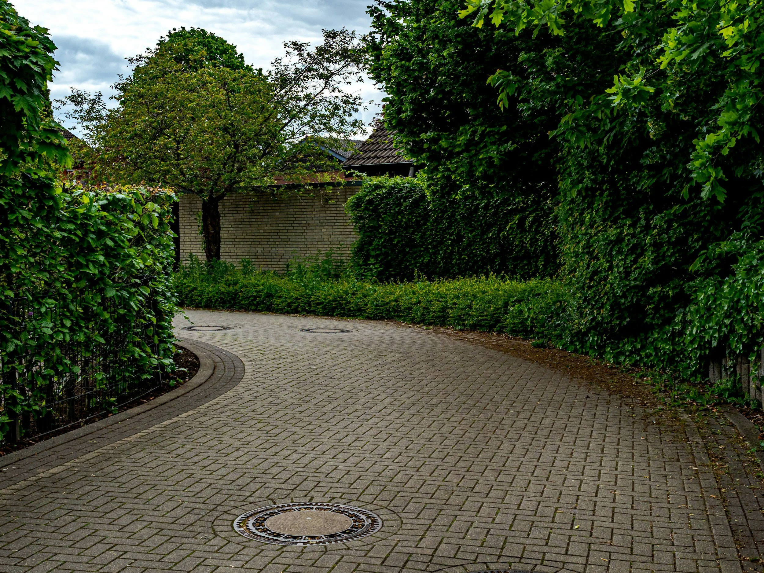 Ein Fußweg in einer grünen Umgebung mit Bäumen und Hecken, umgeben von Zäunen und einer Hausmauer im Hintergrund.
