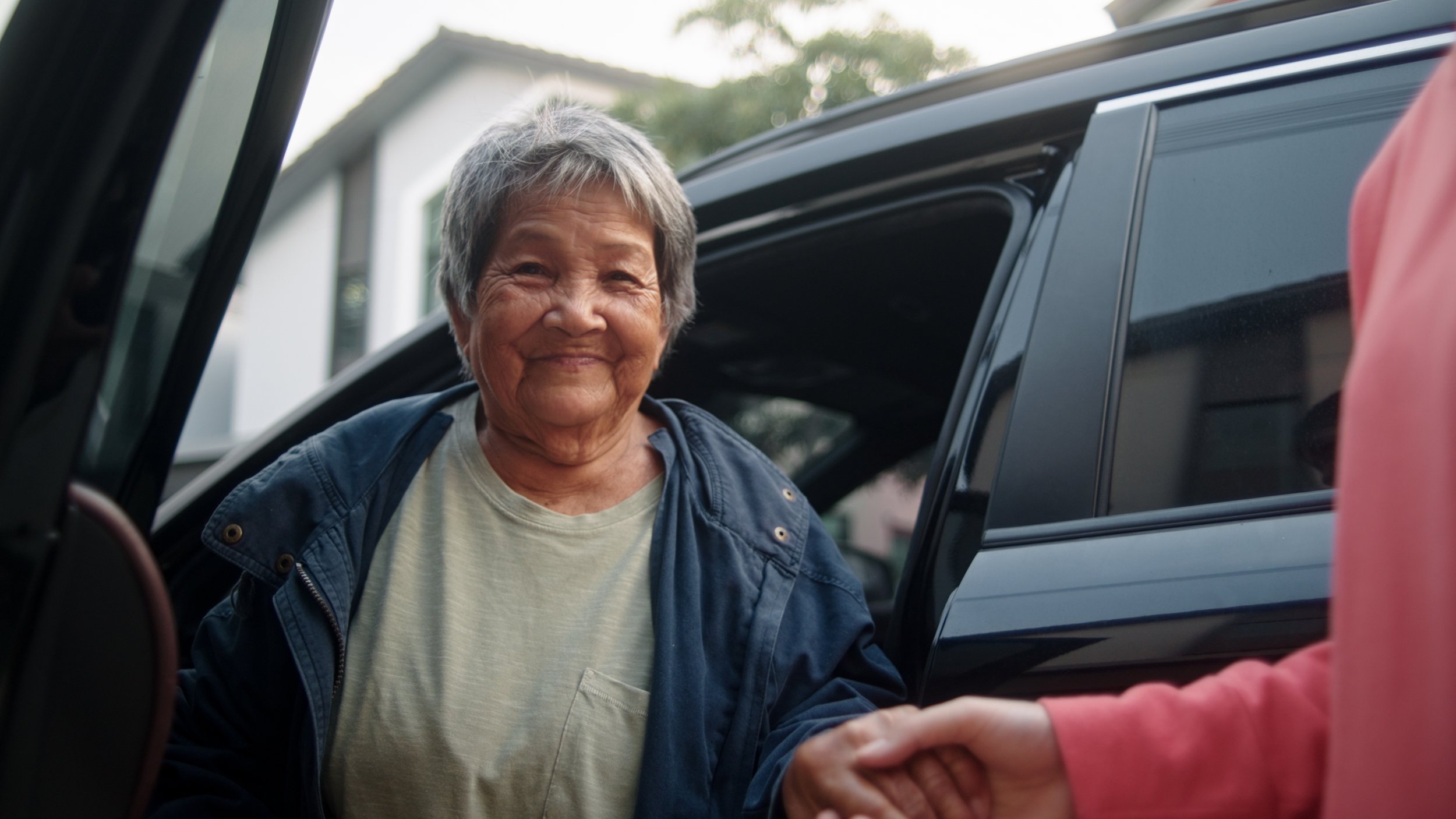 An elderly woman with short gray hair smiling as she accepts a handshake from a person outside a car. The woman is wearing a light-colored shirt and a blue jacket, and the setting appears to be outdoors near a house.