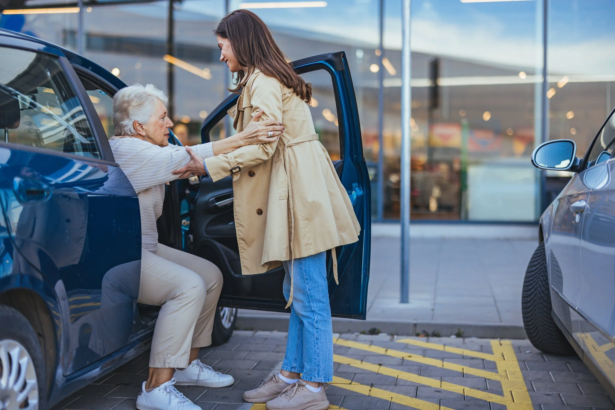 A woman and an elderly woman smiling while greeting each other outside a modern glass building, with cars parked nearby. The elderly woman is seated in a car, and the younger woman is leaning in to greet her with a hug. It appears to be a warm and joyful moment.
