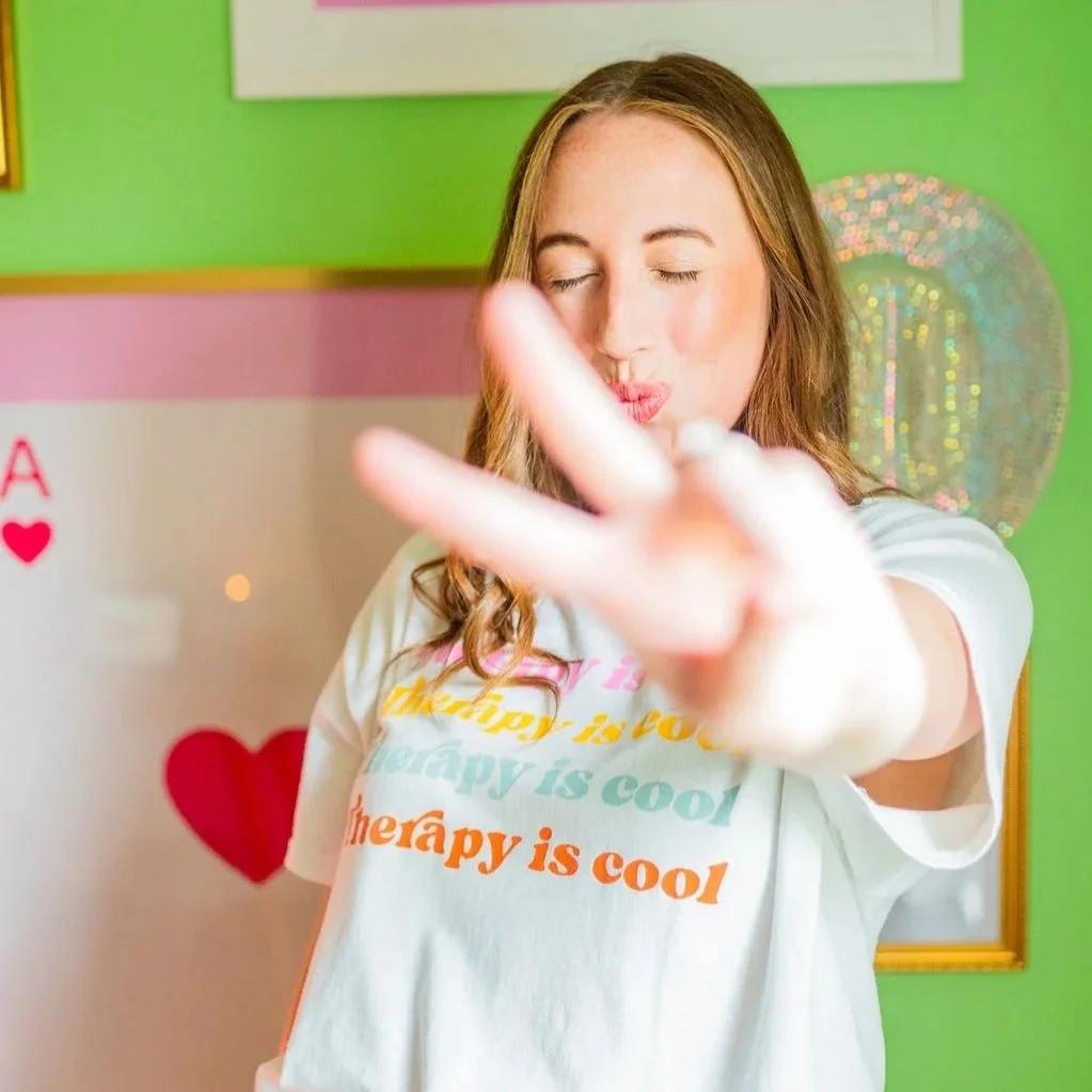 A young woman with long, wavy brown hair making a peace sign gesture toward the camera. She is wearing a white T-shirt with colorful text that reads 'therapy is cool.' The background includes a pink and green wall with a colorful balloon and a whiteb