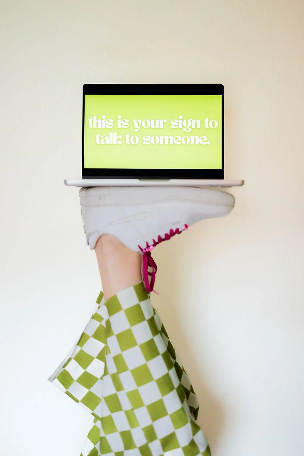 A person balancing on one foot wearing a white sneaker with pink laces and green checkered pants, holding a laptop with a yellow screen that has white text reading 'this is your sign to talk to someone,' against a plain light background.