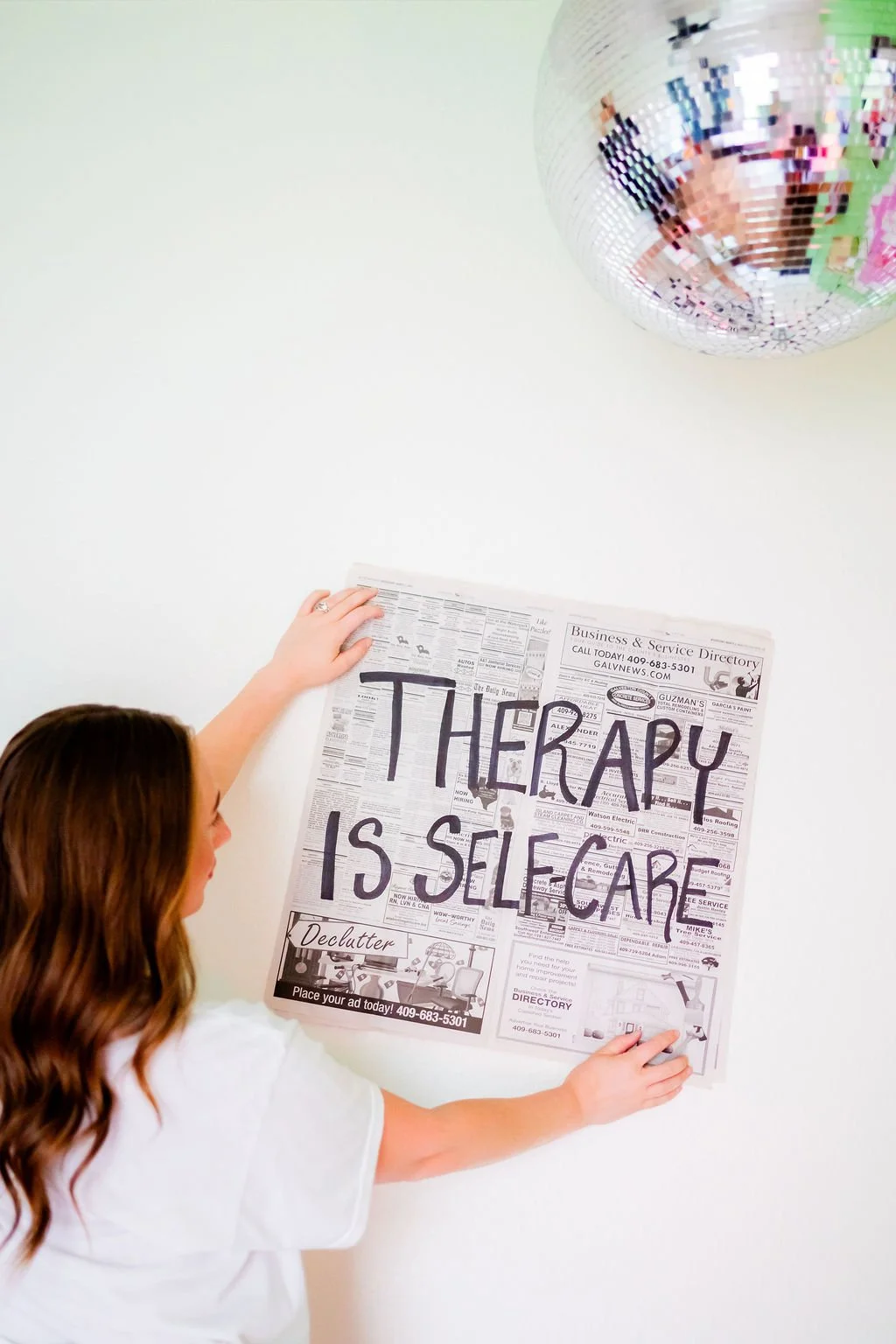 Person hanging a newspaper sign that reads 'THERAPY IS SELF CARE' on a white wall, with a reflective disco ball hanging above.