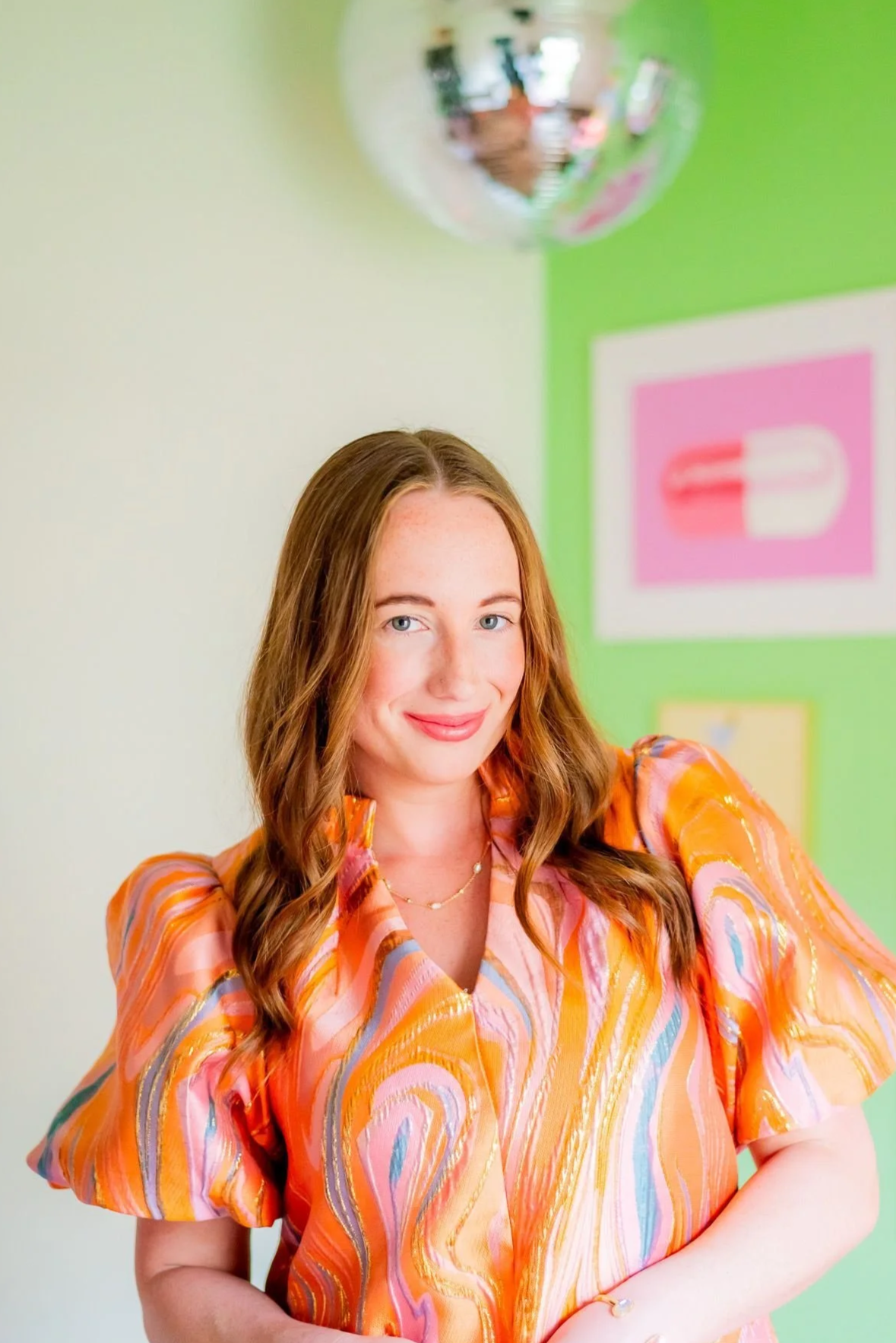 Therapist in Texas standing in a colorful counseling office with a green wall, framed artwork, a disco ball, & an orange dress, representing a welcoming virtual therapy practice.