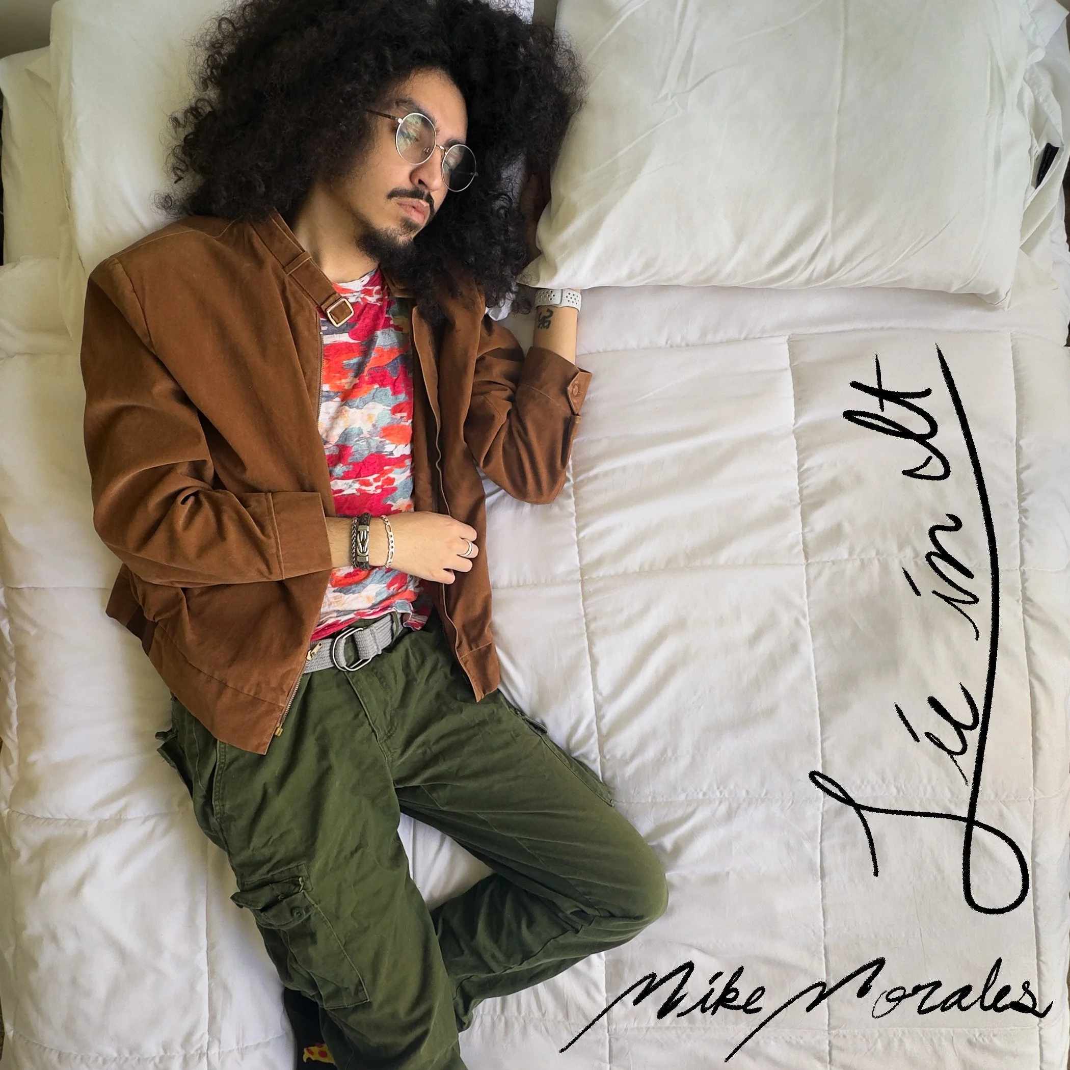 An album cover with a man with curly hair, wearing glasses, a brown jacket, and green pants sleeping on a white bed with white pillows. Cursive text on the bed reads 'Lie in It' with his name below: 'Mike Morales'.
