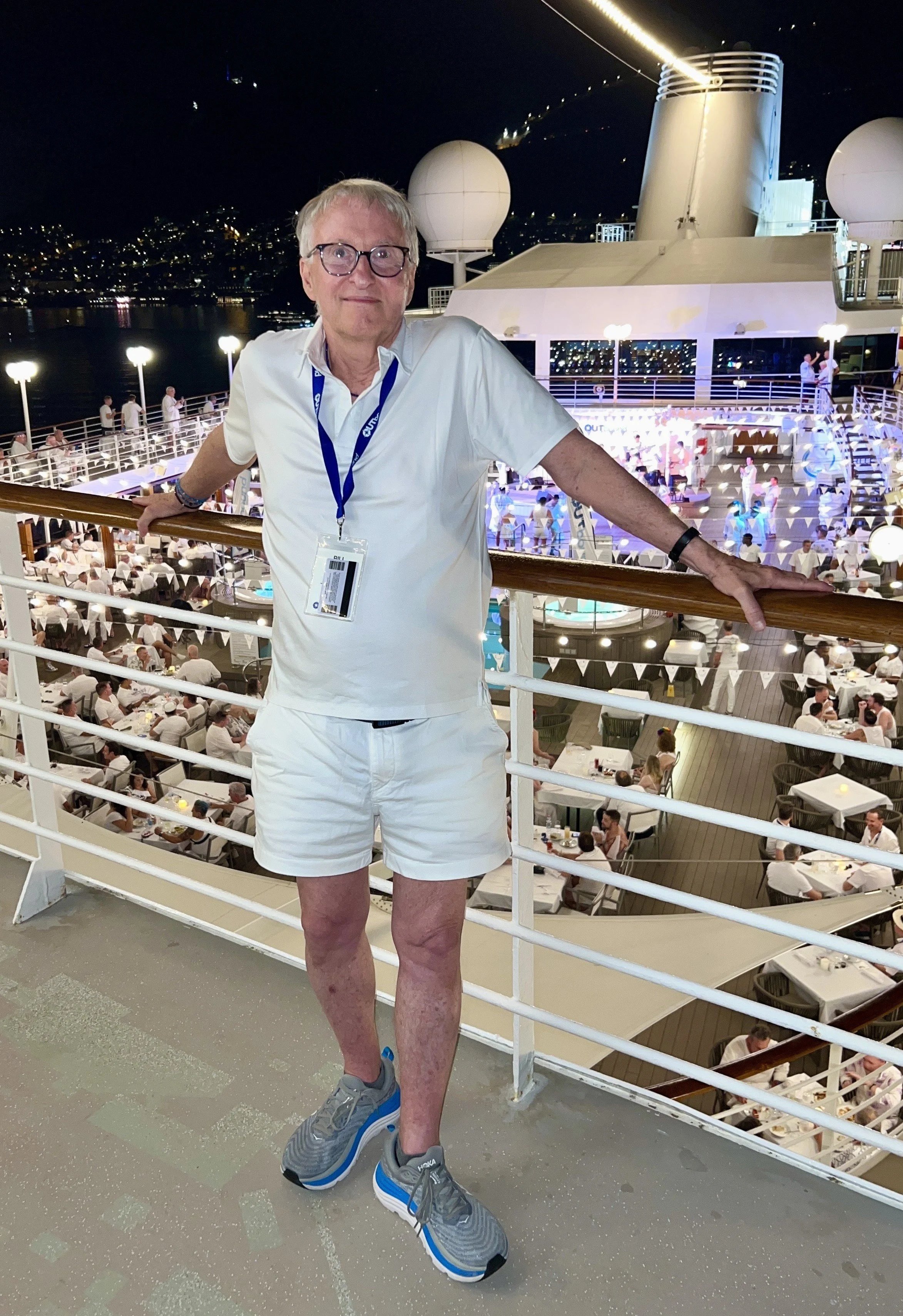 A man wearing glasses, a white shirt, white shorts, and gray sneakers standing on a deck at night with a large cruise ship in the background, illuminated with artificial lighting and city lights in the distance.