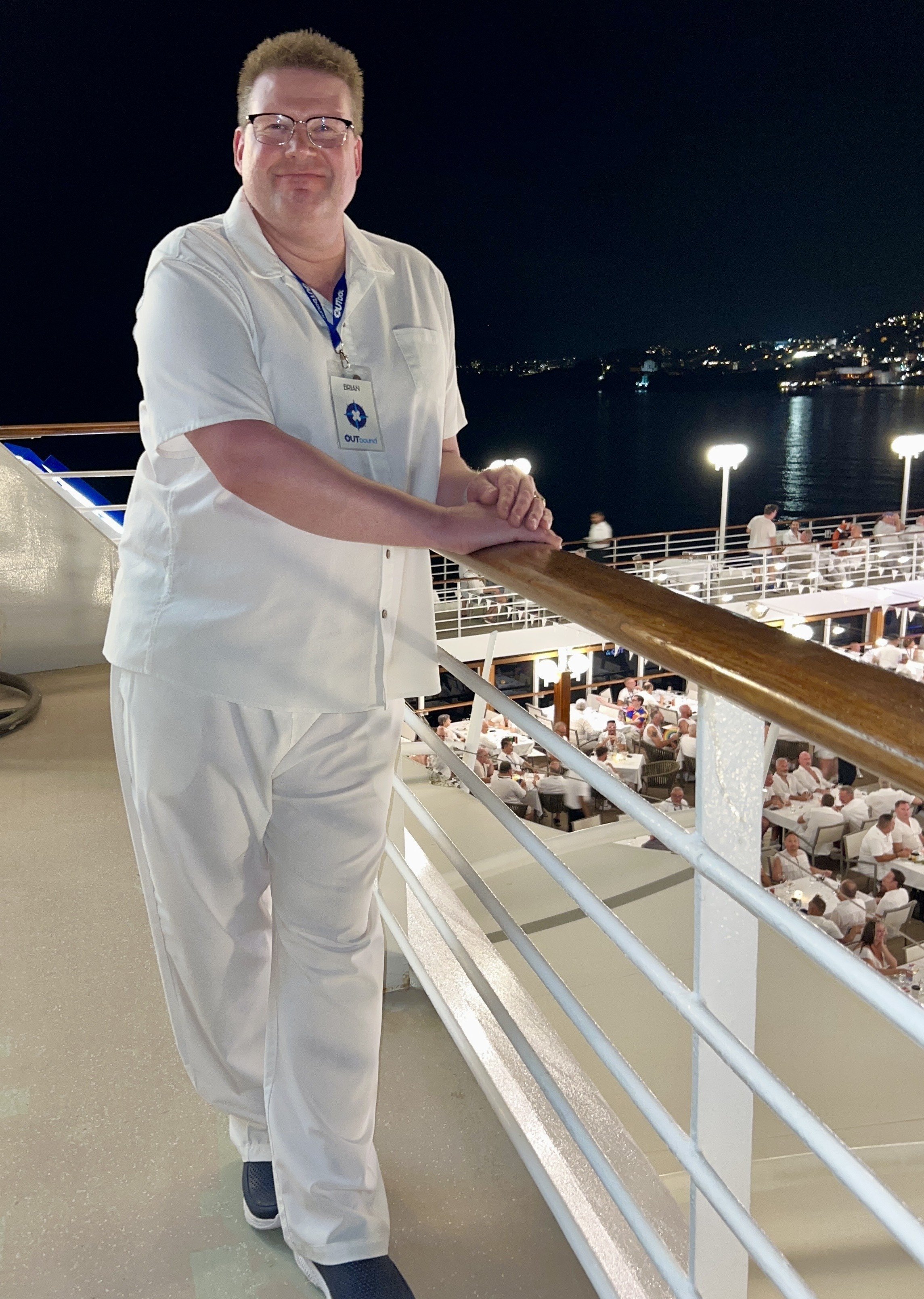 A man wearing a white shirt and white pants with a cruise ship logo around his neck, standing on the deck of a cruise ship at night, with the ocean and a city skyline in the distance, overlooking a crowded dining area below.