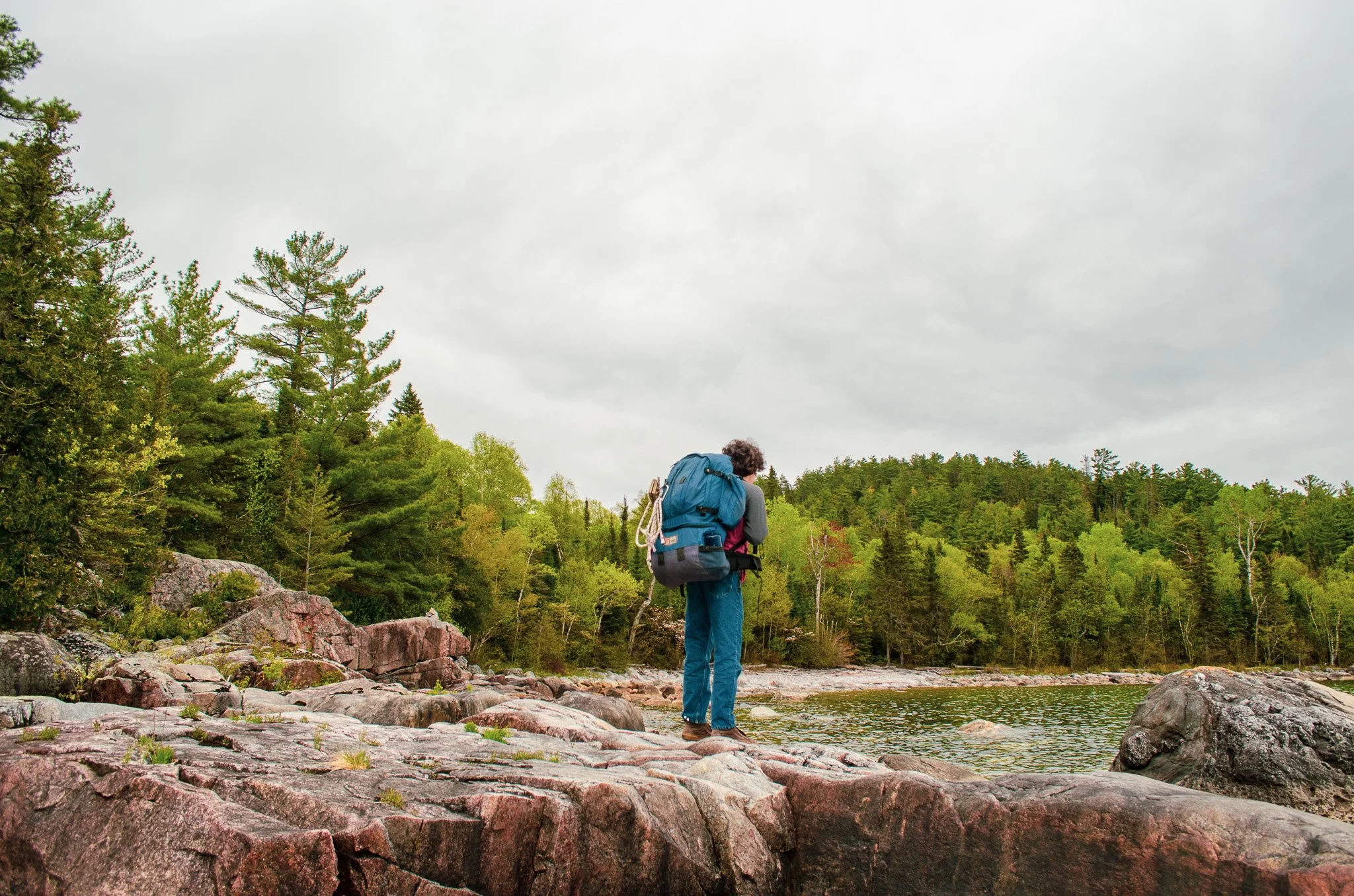 Hiker standing by a riverbank, surrounded by trees, with mountains in the background under a cloudy sky.