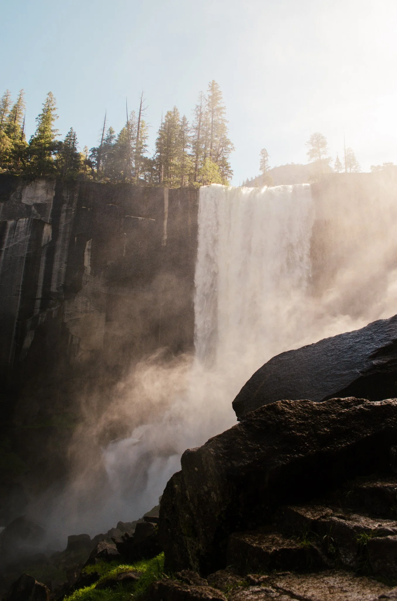 View of a large waterfall cascading down a rocky cliff with a forest of tall pine trees at the top, mist rising at the base, and sunlight illuminating the scene.