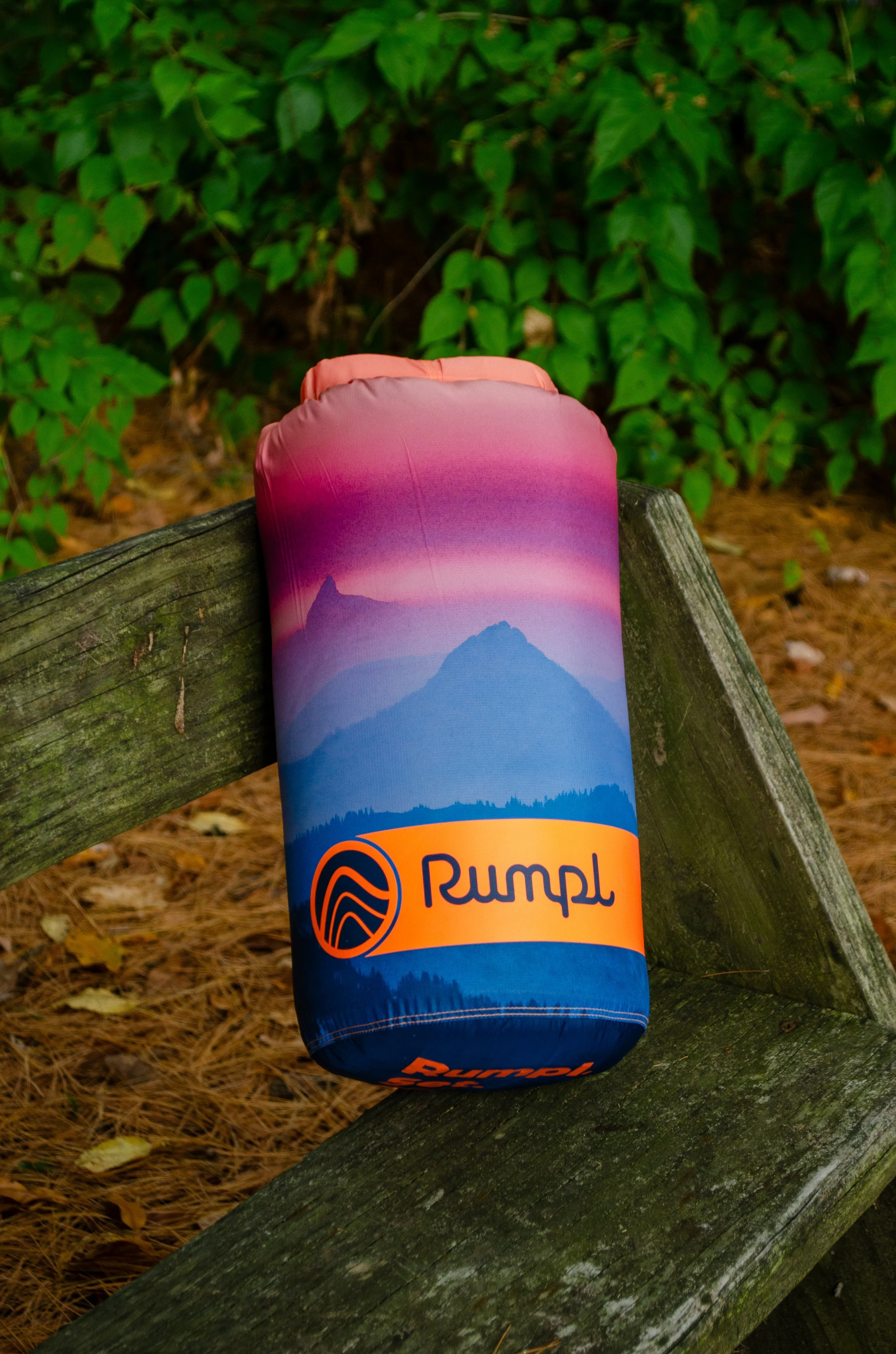 A rolled-up rumpl blanket with mountains and a sunset or sunrise scene, placed on a wooden bench outdoors, surrounded by green foliage and pine needles.
