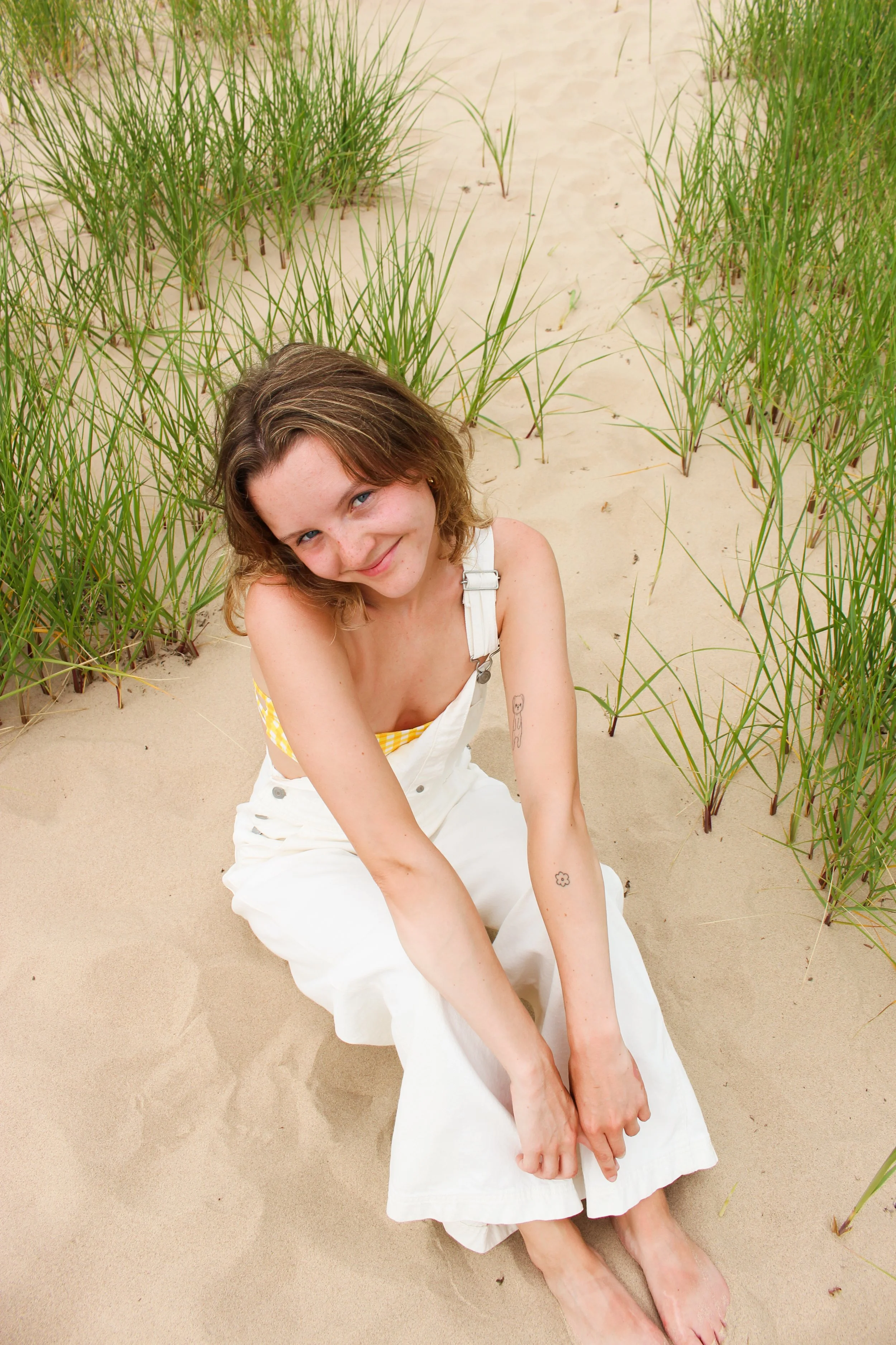 A young woman with light brown hair and blue eyes is sitting on a sandy beach surrounded by tall green beach grasses, smiling at the camera.