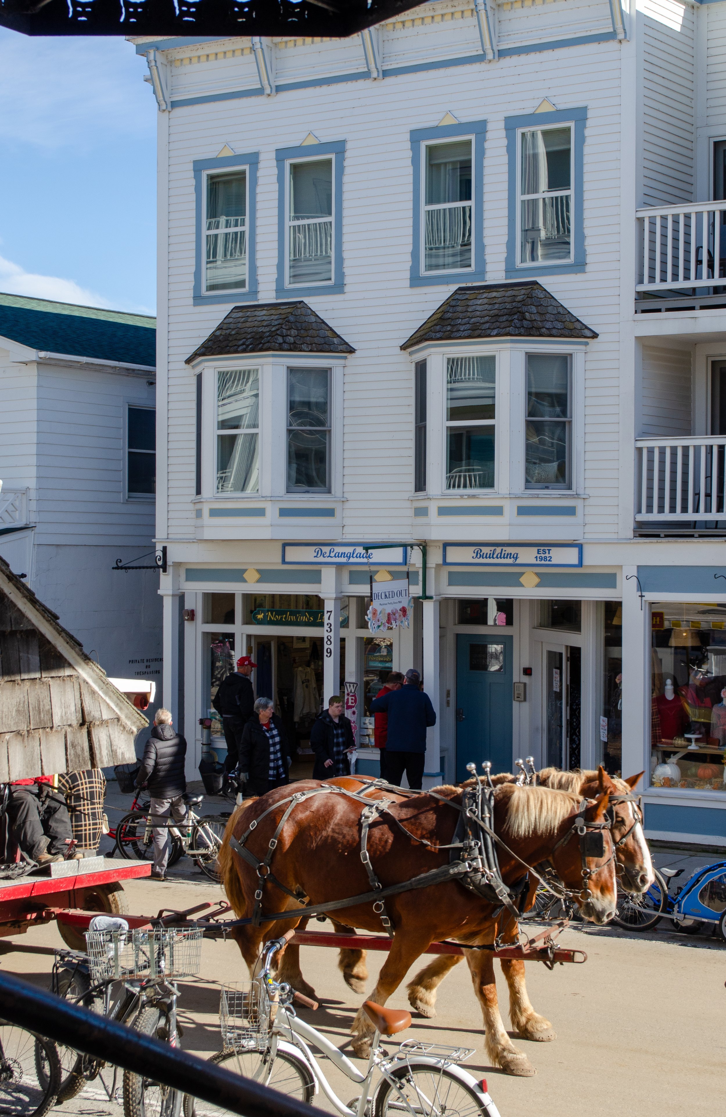 A busy street scene in front of a white multi-story building with blue trim. There are people walking, bicycles parked, and a team of horses pulling a carriage. The building has signs that read 'DeLanglade Building' and 'Northwinds'.