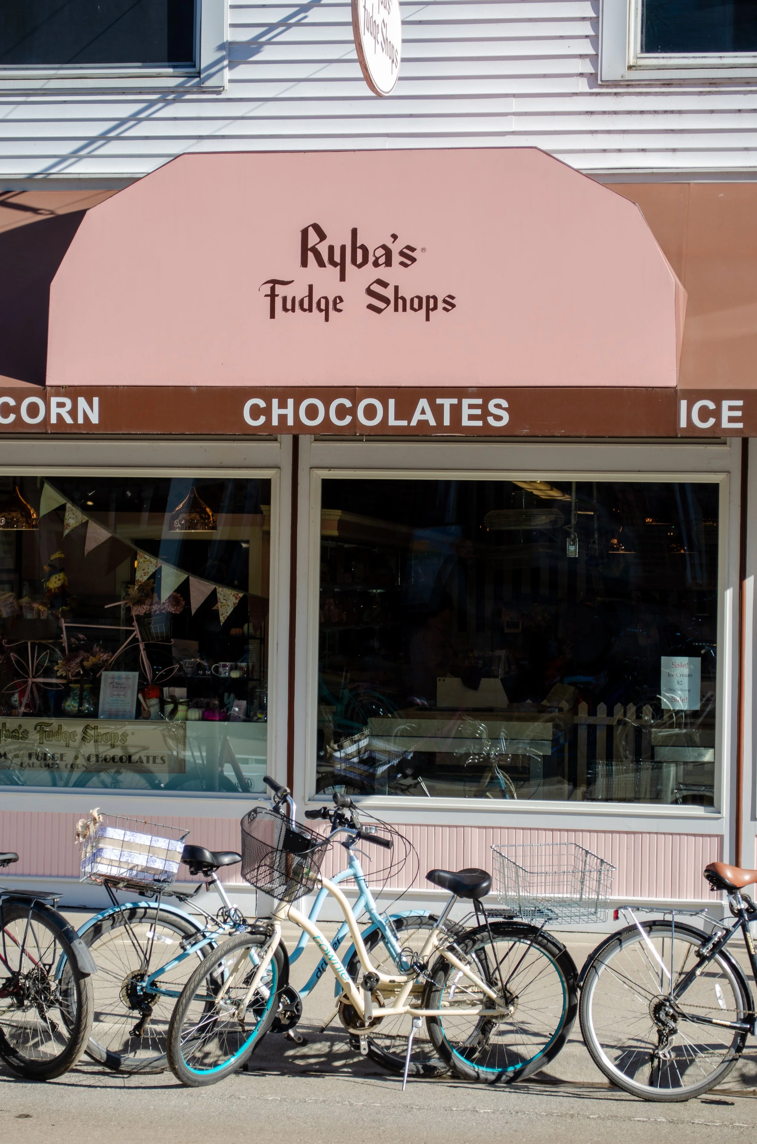  storefront with a pink awning reading 'Ryba's Fudge Shops' and a brown stripe with words 'CORN', 'CHOCOLATES', and 'ICE'. In front, bicycles are parked on the sidewalk with a building visible inside.