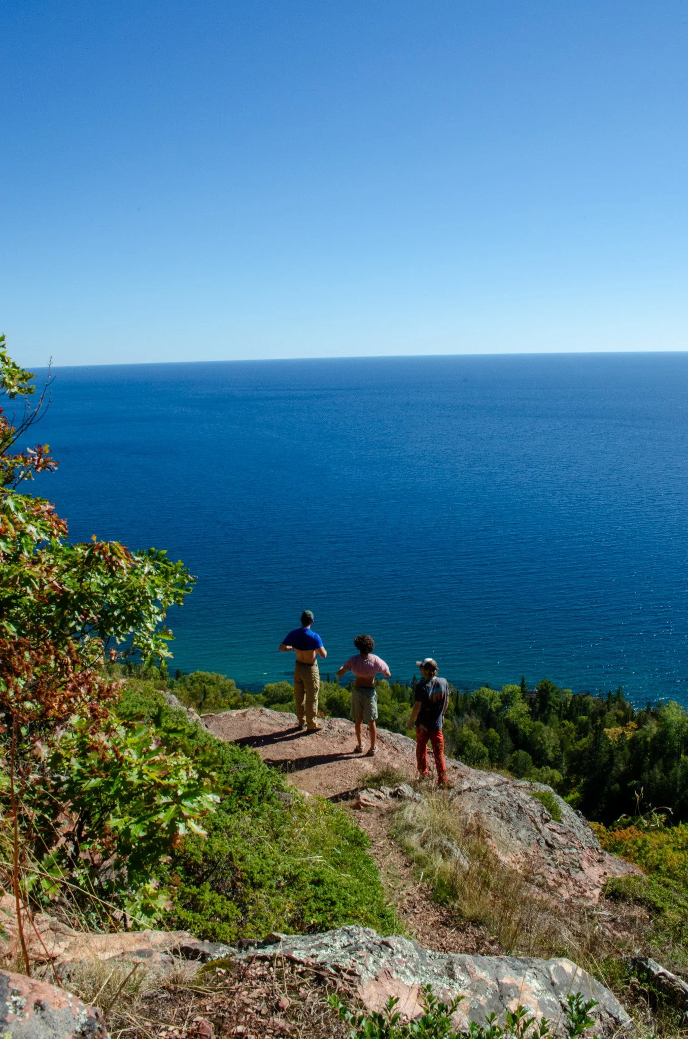 Three people standing on a cliff overlooking a large body of water with a clear blue sky, some greenery, and trees in the background.