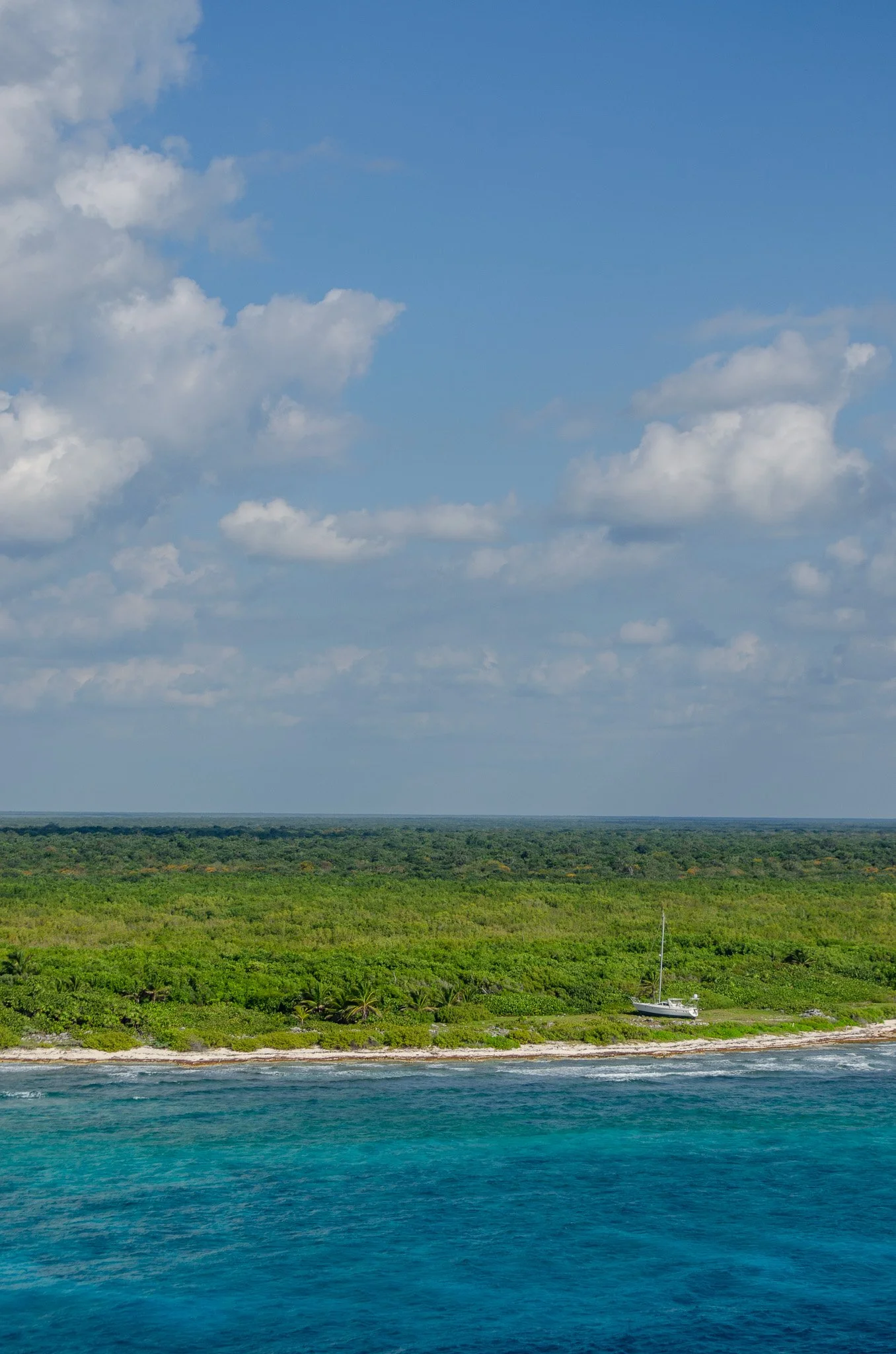 Tropical landscape with green forest, a small boat near the shore, and a vast blue ocean in the foreground. The sky is partly cloudy with fluffy white clouds.