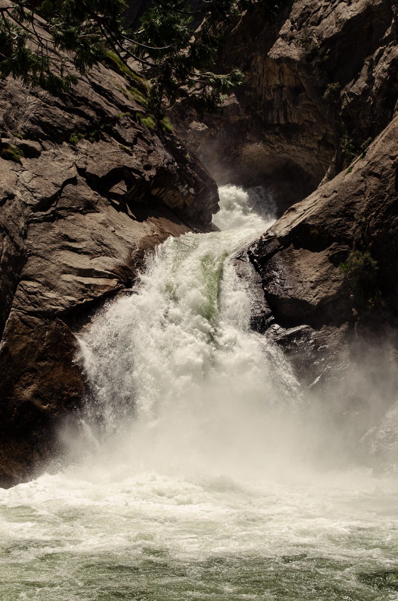 View of a canyon with a river rushing through, surrounded by tall rocky cliffs and sparse greenery.