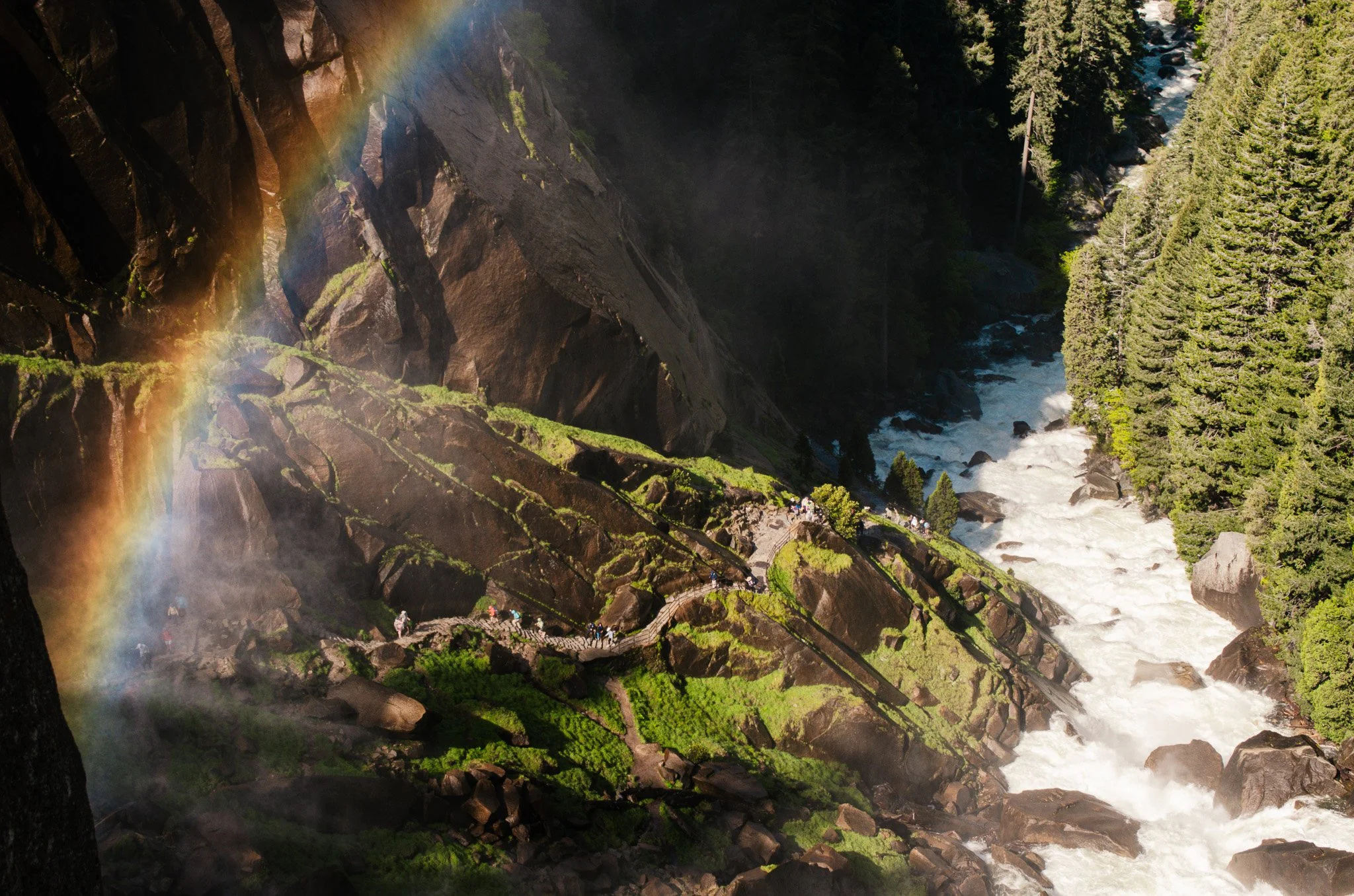 A scenic view of a waterfall with a rainbow, surrounded by tall trees and rocky terrain, with a pathway and people in the distance.