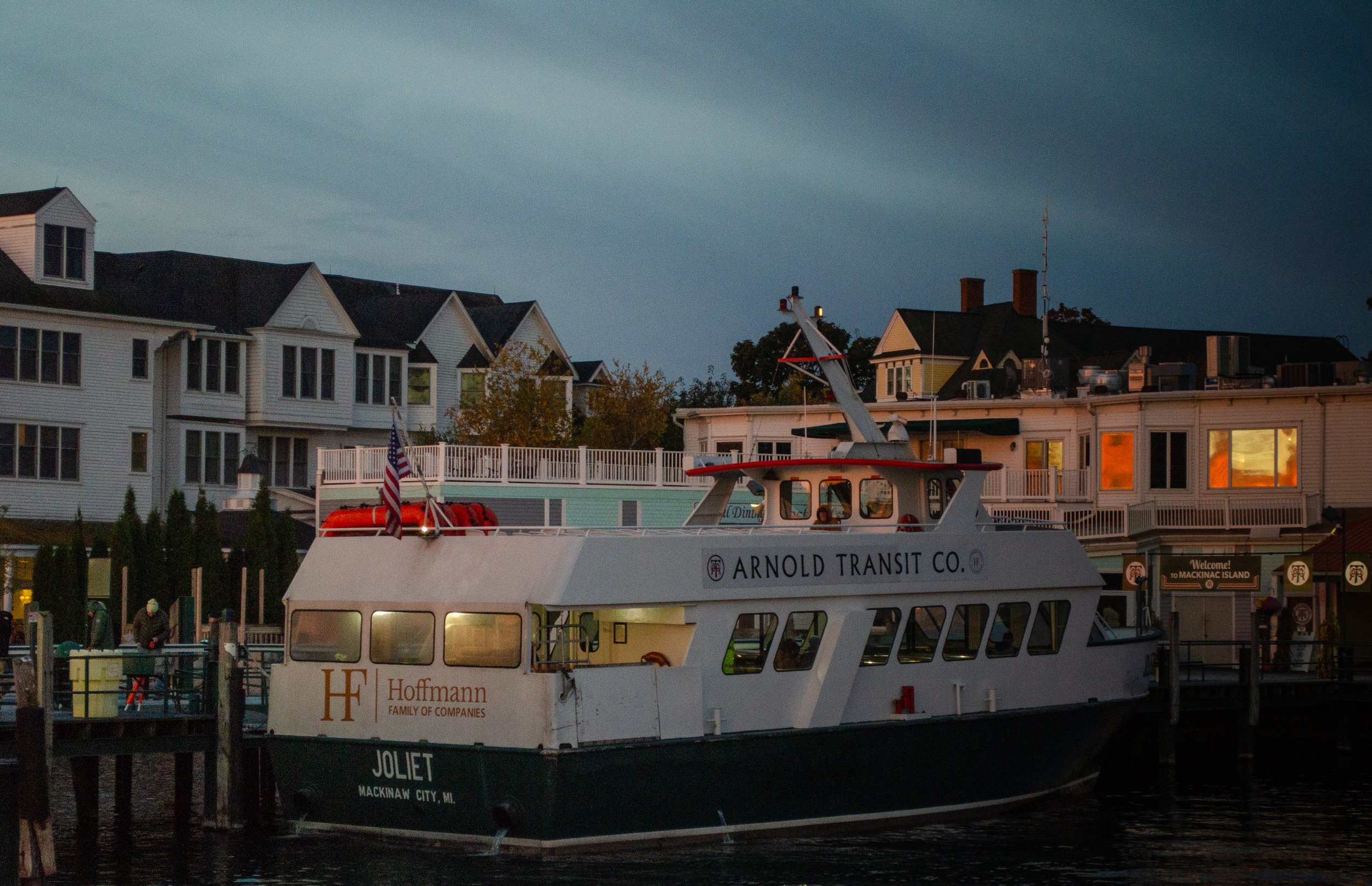 A boat named Arnold Transit Co. docked at a harbor with houses and trees in the background during dusk, with dark clouds overhead.