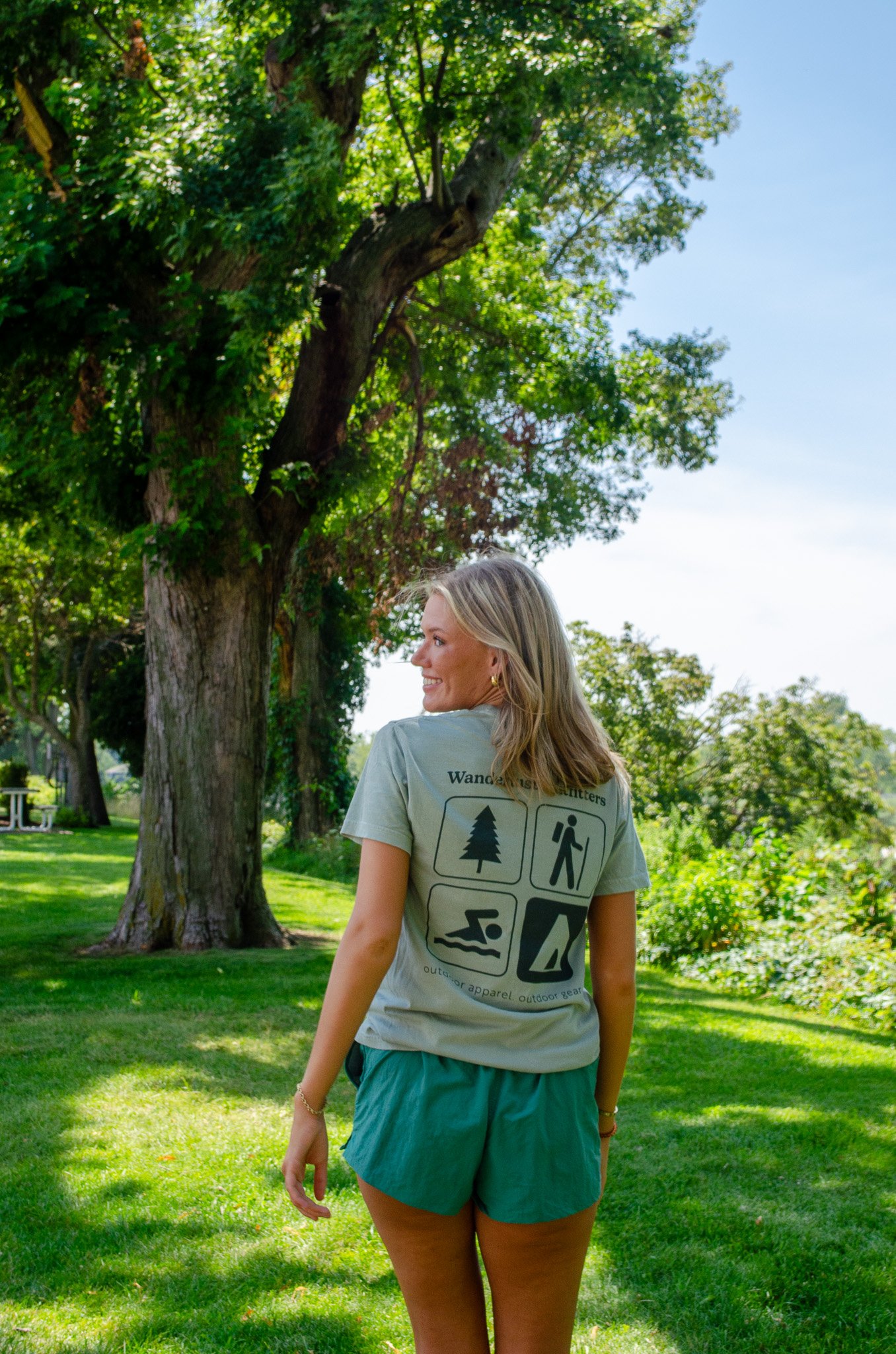 A woman smiling in a park with green grass and trees, wearing a tan T-shirt and teal shorts.