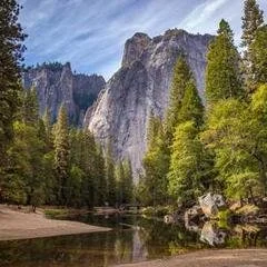 A scenic landscape of a river flowing through a forested area with tall mountains in the background under a blue sky.