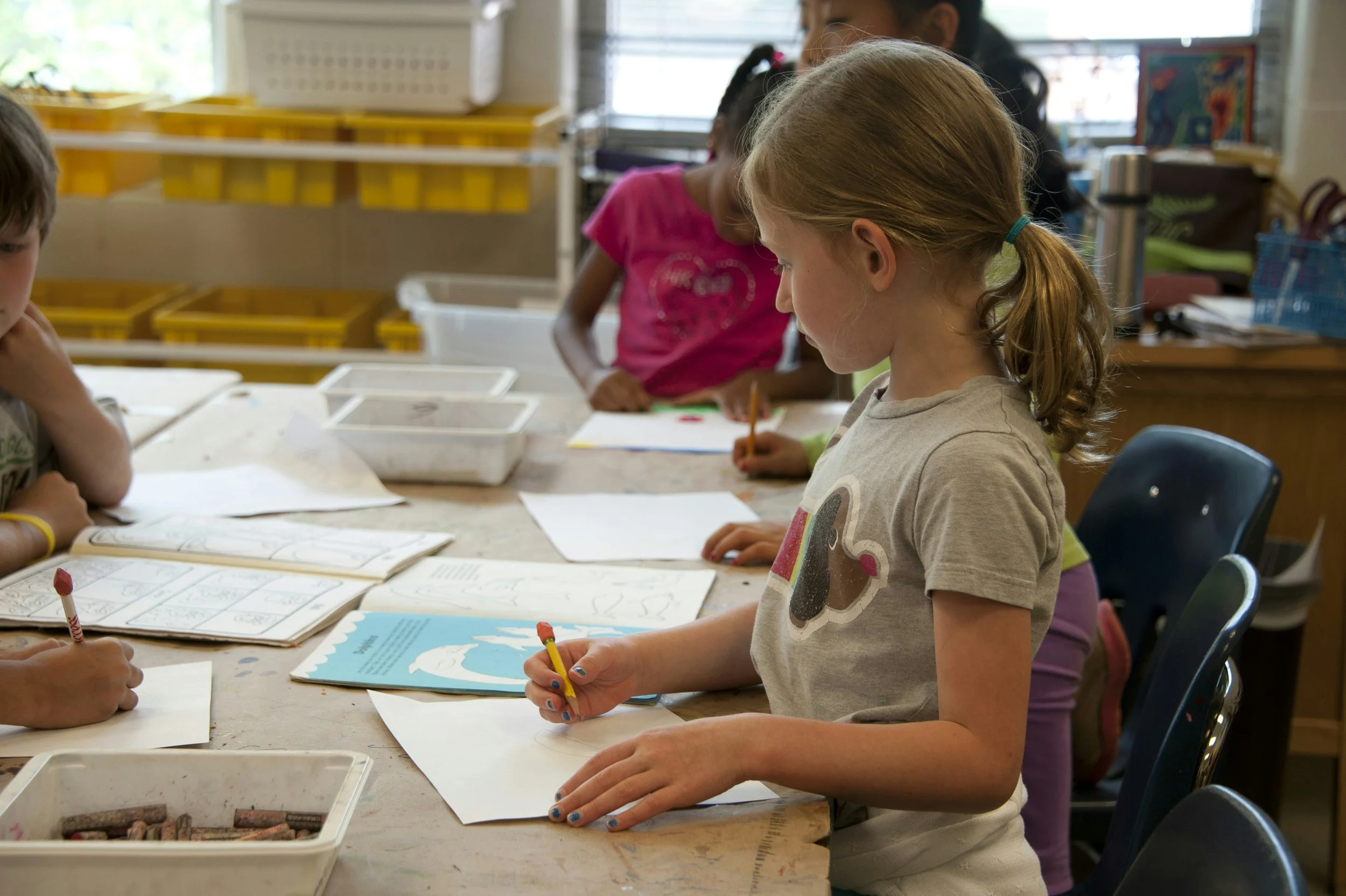 Children sitting at a table engaging in arts and crafts activities in a classroom, with papers, crayons, and drawings on the table.