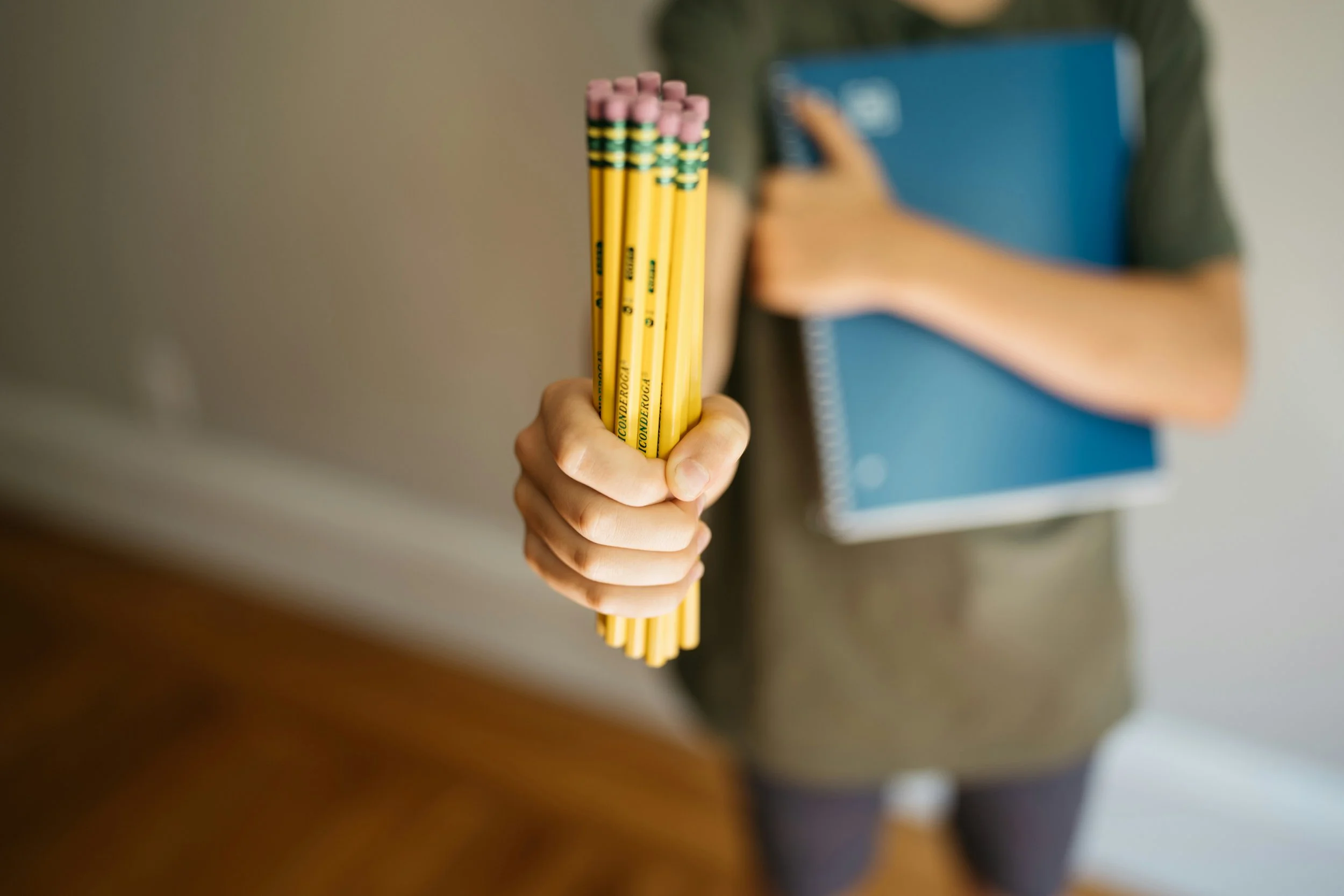 Close-up of a hand holding a bundle of yellow pencils with pink erasers and green and yellow stripes, with a person in the background holding a blue notebook.