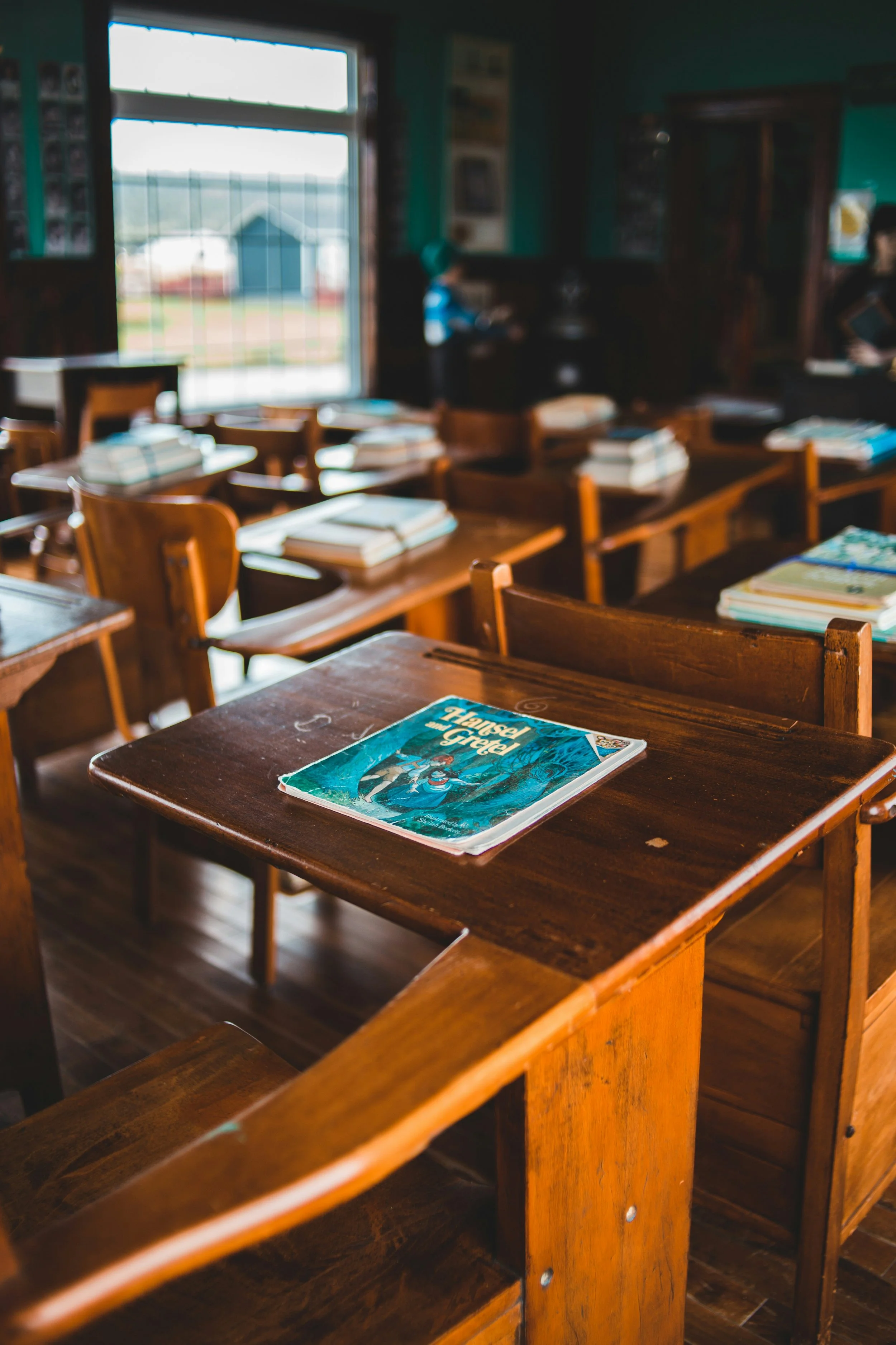 A classroom with multiple wooden desks and chairs, books stacked on each desk and a large window in the background.