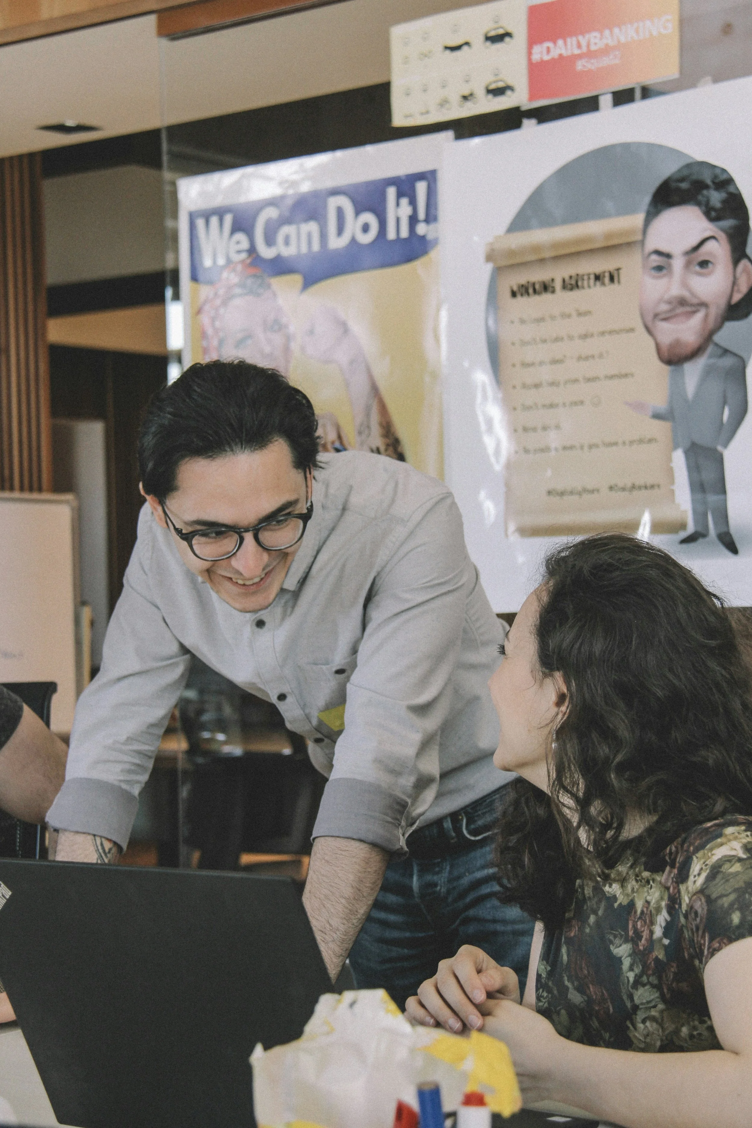 A group of people working together at a table in an office or cafe. One person is smiling and leaning over, while two others are seated, one with long dark hair and a floral blouse, and another with glasses and dark hair. There are posters on the wall, including a motivational poster that says 'We Can Do It!' and a cartoon figure. The group appears engaged in a collaborative activity.