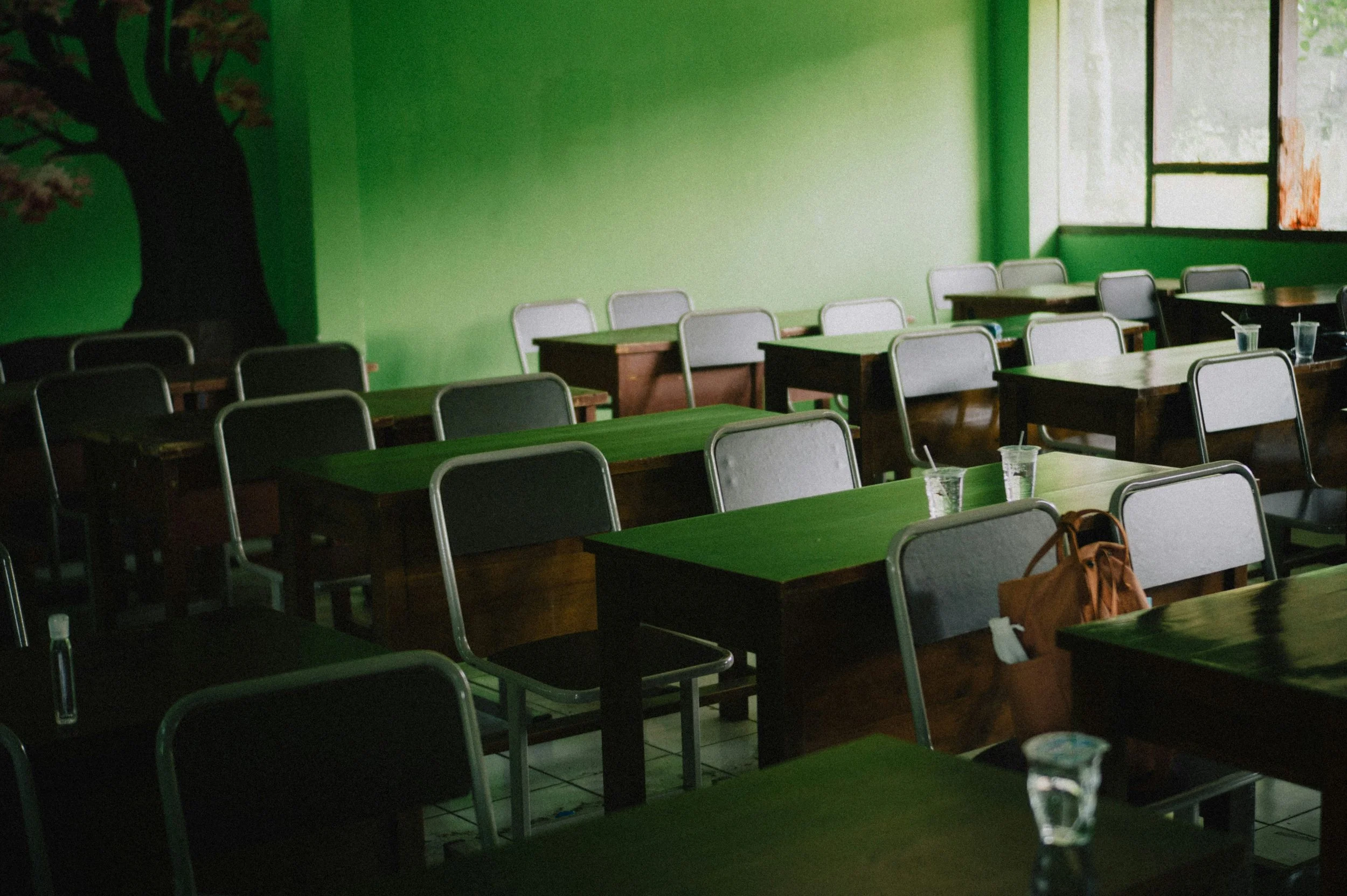 Empty classroom with green walls, multiple desks and chairs, some drinks on the tables, large windows letting in natural light.