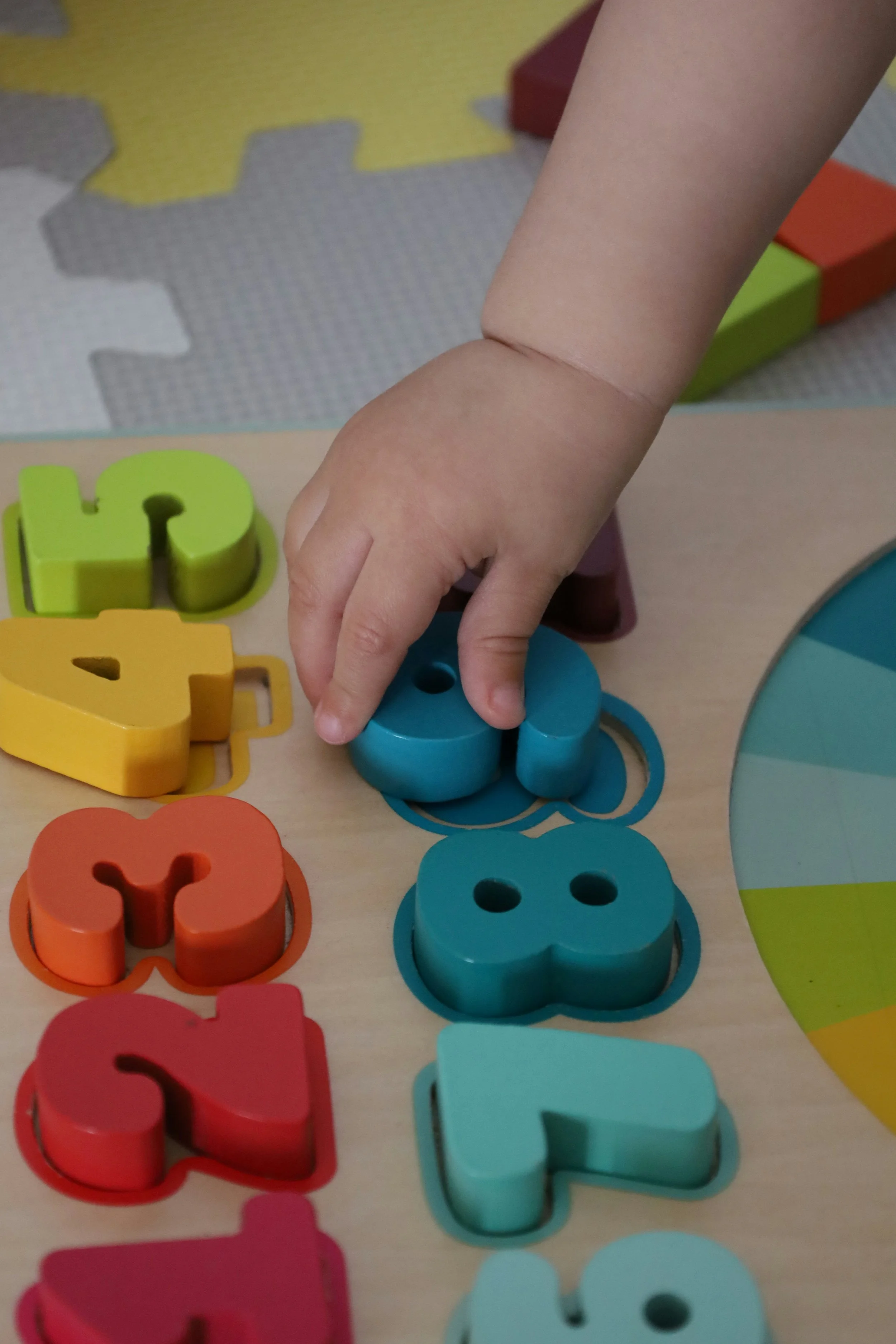 Child's hand placing a blue wooden block with two holes into a corresponding cutout on a wooden activity board. Colorful shaped blocks are nearby.