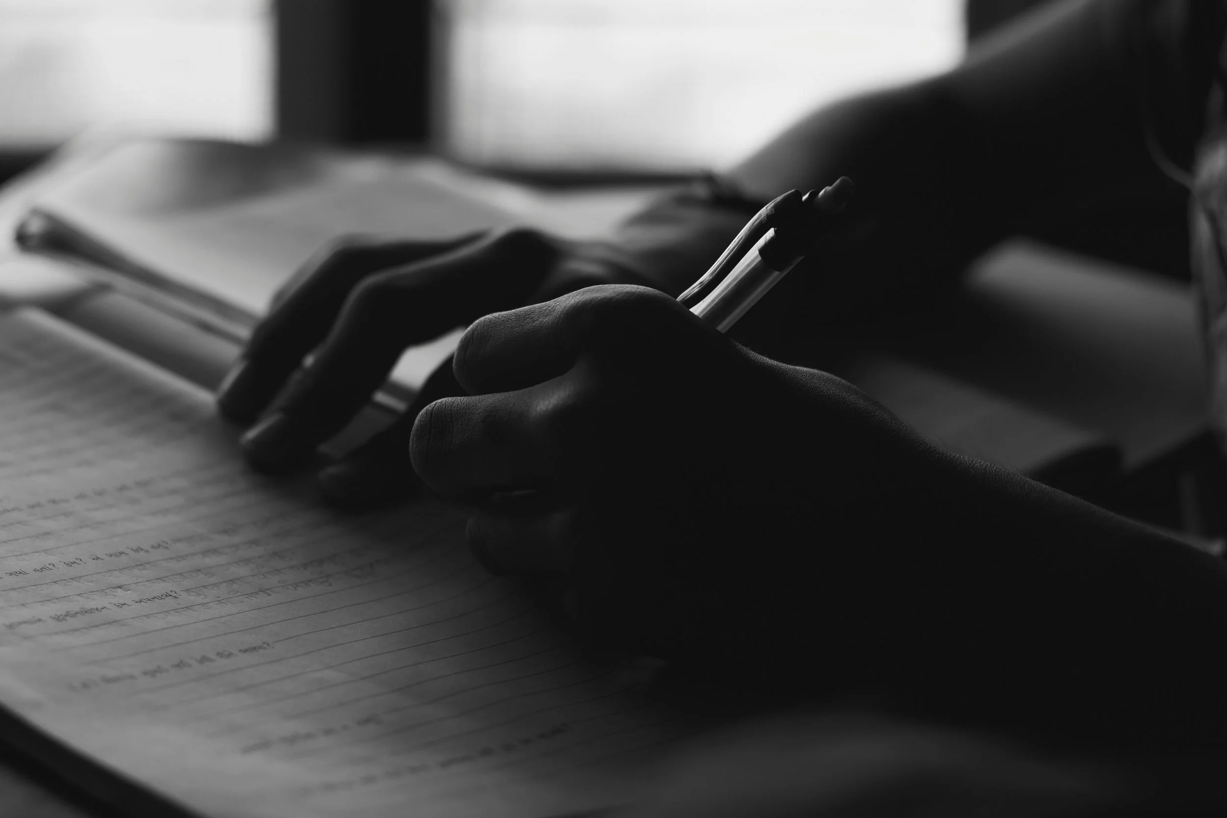 Close-up of a person's hand holding a pen, writing on lined paper, with an open notebook nearby, in black and white.
