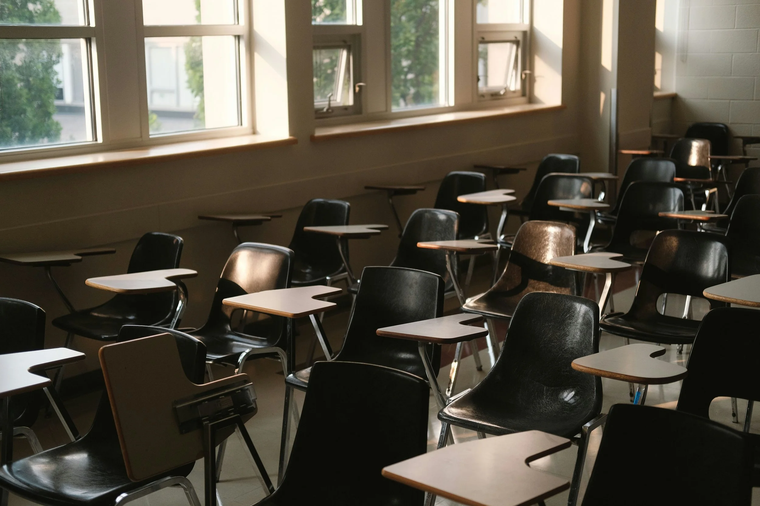 Empty classroom with several black chairs with attached writing surfaces, large windows letting in natural light, and a view of trees outside.