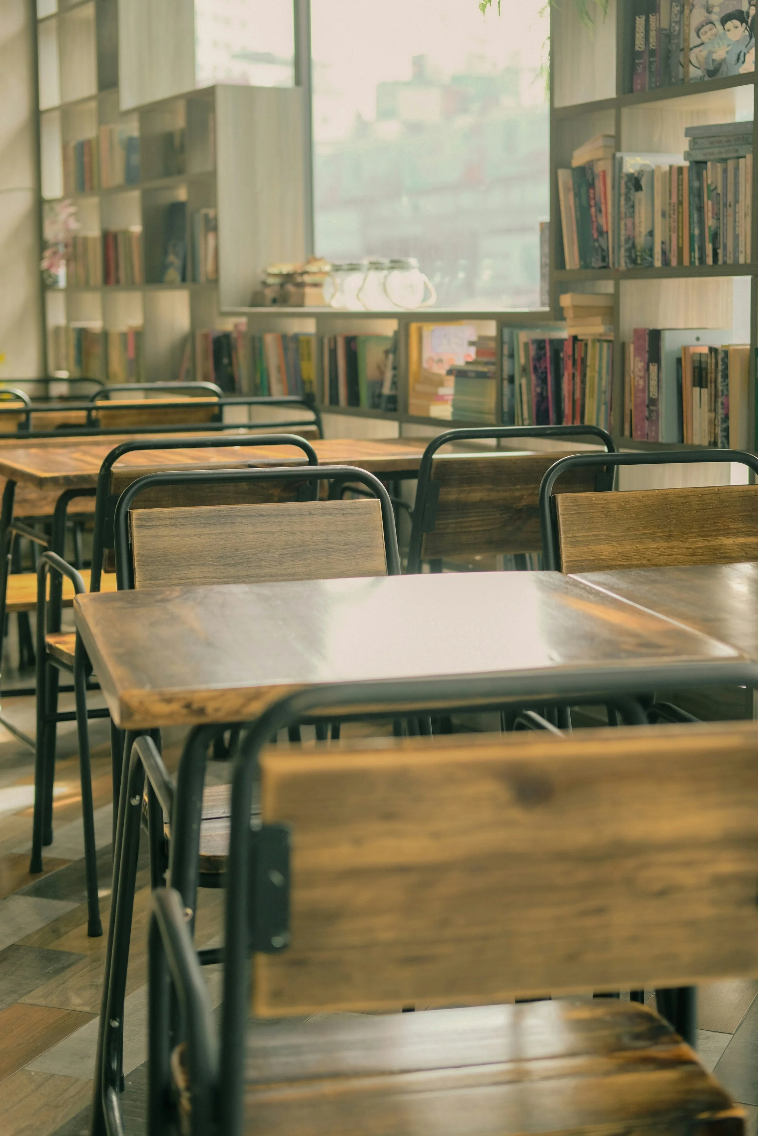 Empty wooden tables and black metal chairs in a cozy, well-lit library or bookstore. Bookshelves filled with colorful books line the walls, with a large window letting in natural sunlight.
