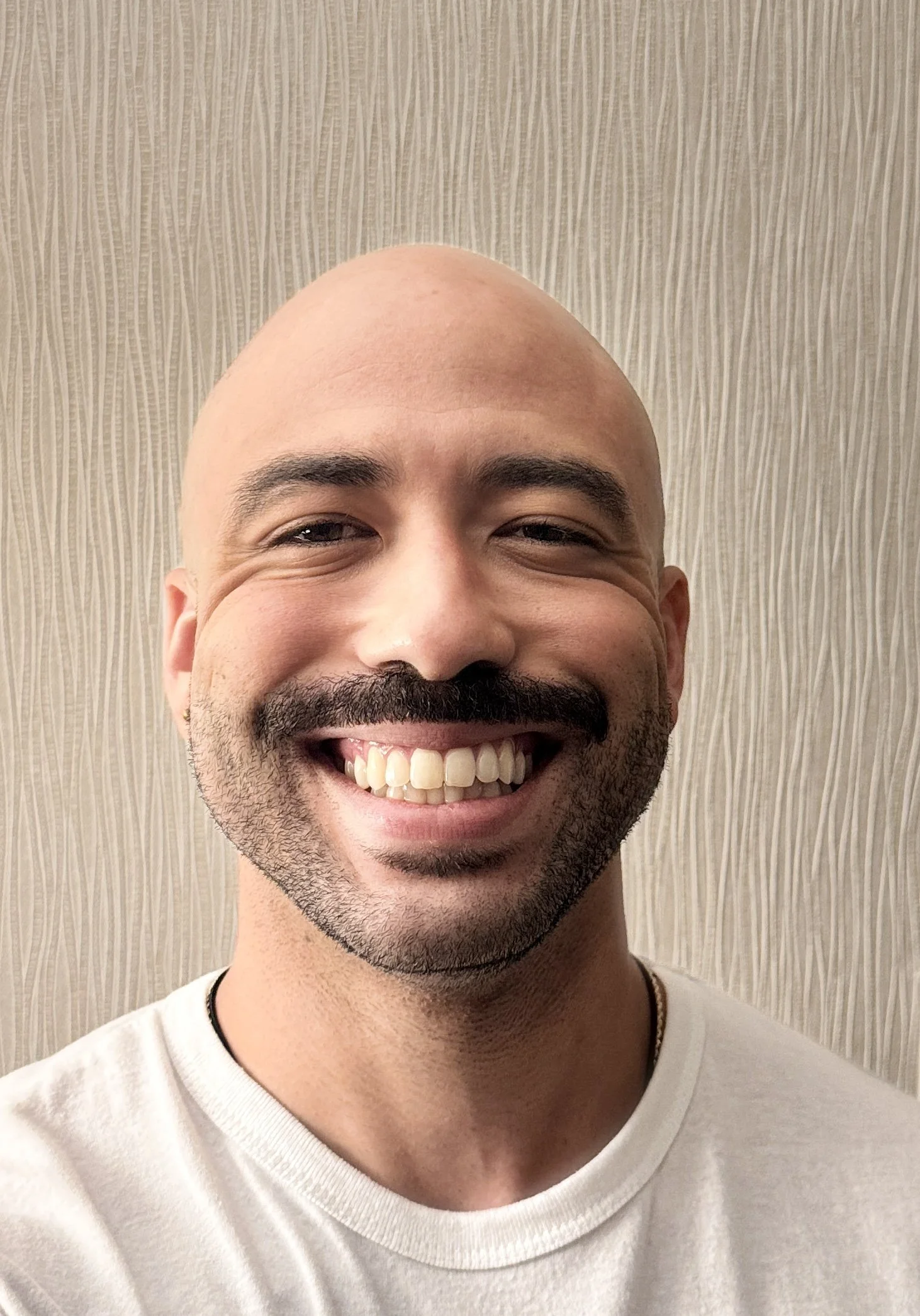 Close-up of a smiling man with a bald head, beard, and mustache, wearing a white shirt, standing in front of a beige textured wall.