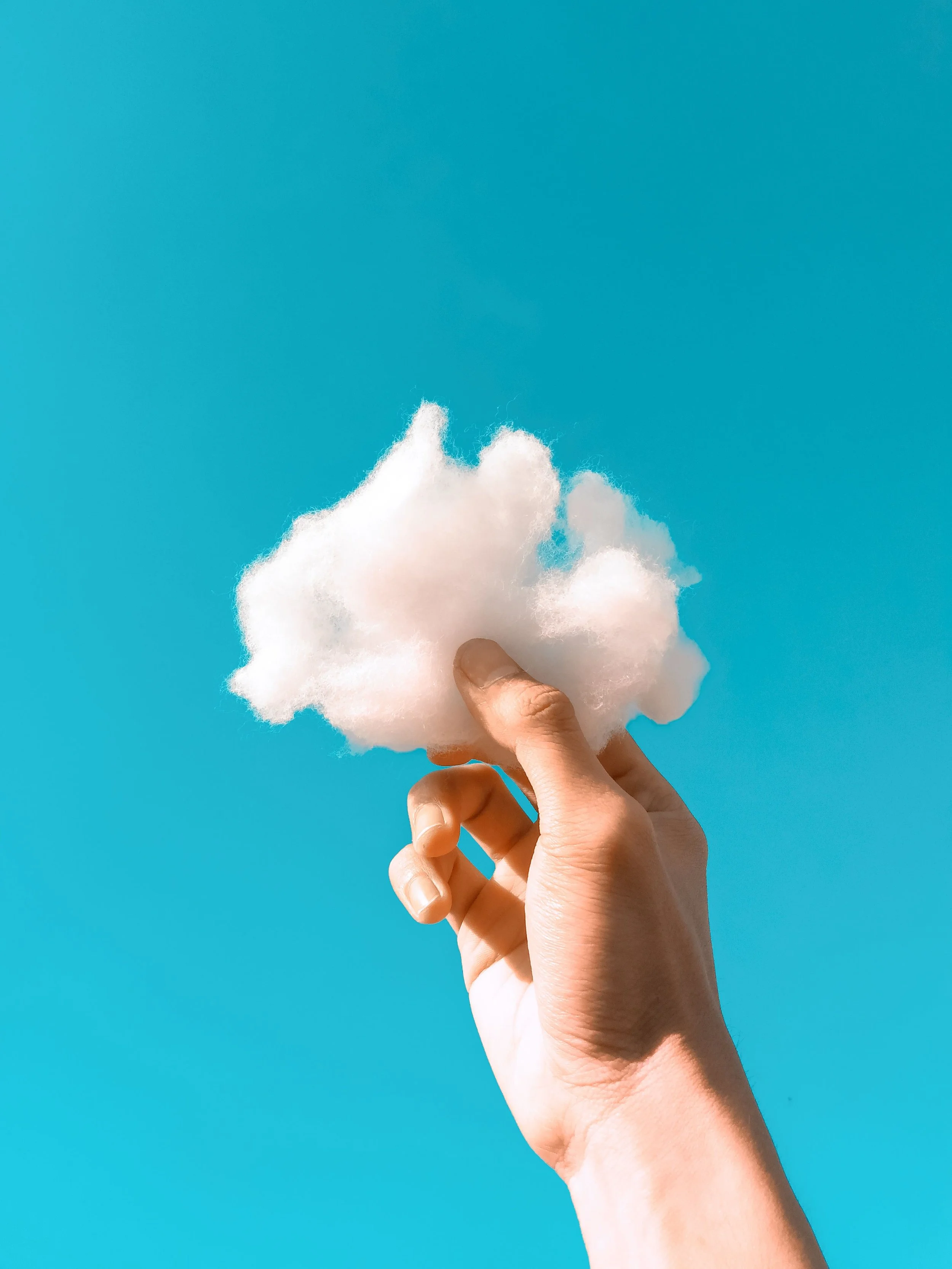 Hand holding a piece of white cotton candy against a bright blue sky.