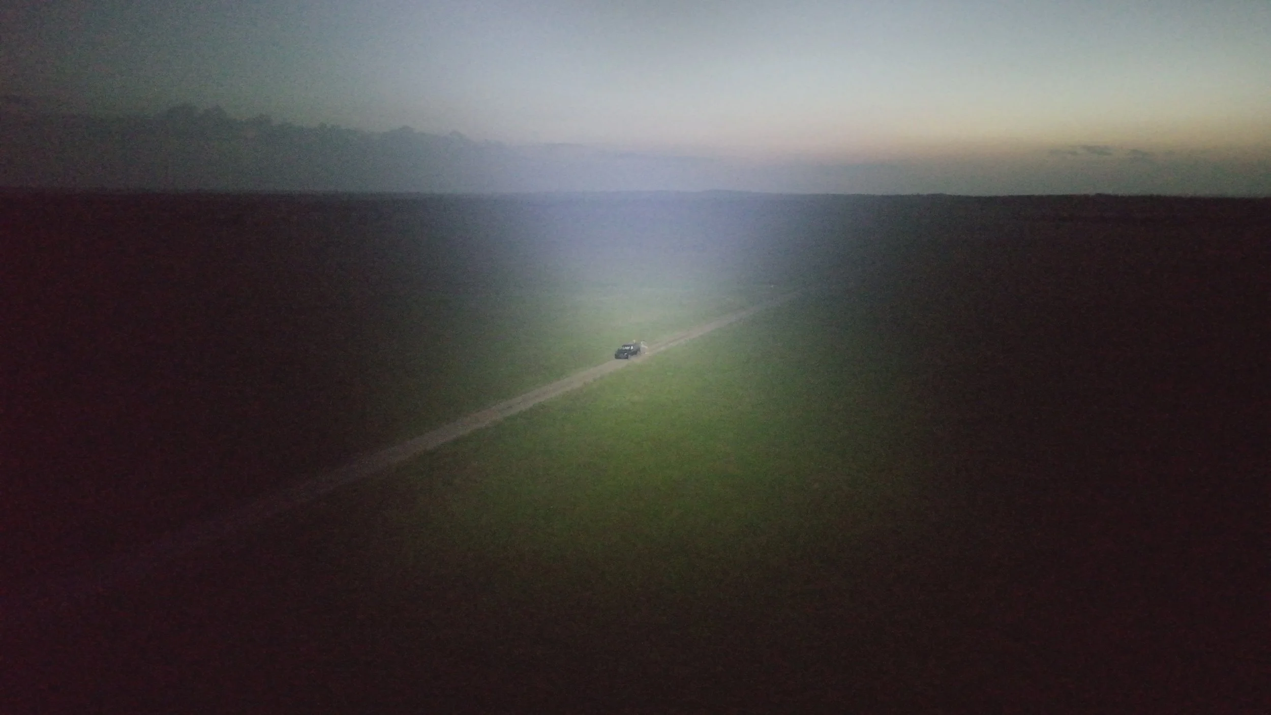 A black vehicle driving on a narrow dirt road through a vast grassy field during dusk or dawn, with dark clouds and a horizon in the background.