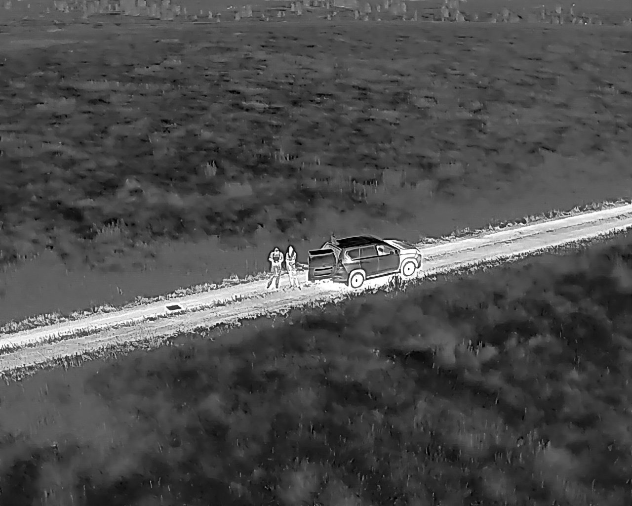 Nighttime infrared image of a rural dirt road with an SUV and two people standing beside it.