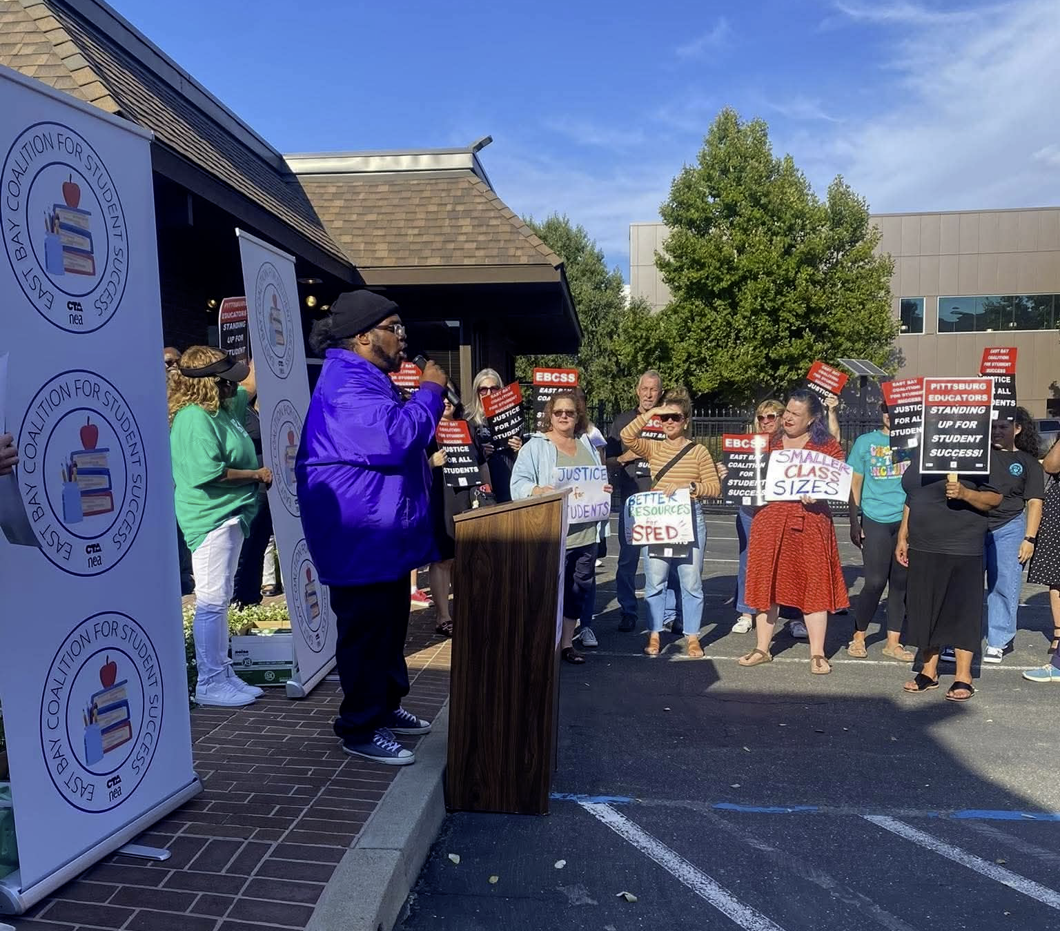 Brandon speaking to a group of people gathered outdoors at a rally for special education resources, with crowd members holding signs calling for justice, better resources, and supporting students, while a man speaks at a podium.