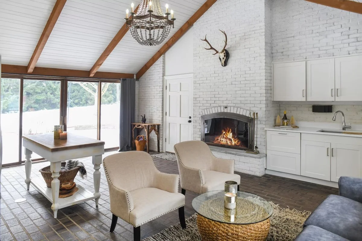 Living room with white brick wall, fireplace, white cabinets, beige chairs, glass coffee table, chandelier, and windowed sliding door leading outside.