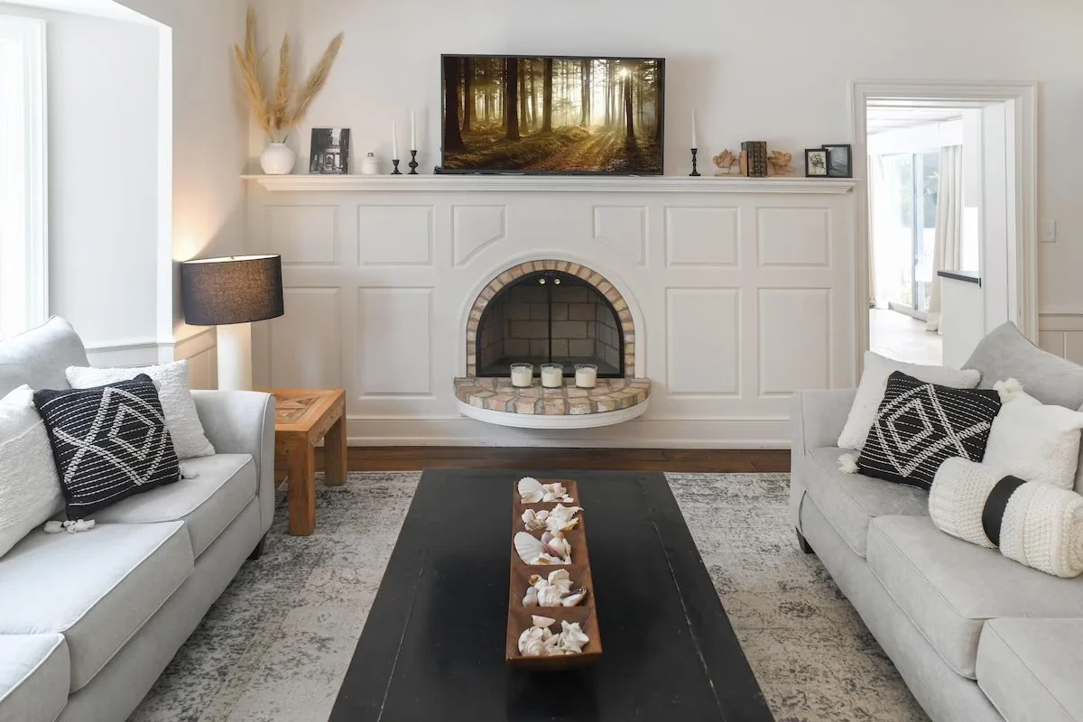 Living room with white sofas, black and white pillows, a black coffee table with seashells, a fireplace with candles, housing a TV displaying a forest scene, and a doorway leading to a bright room.