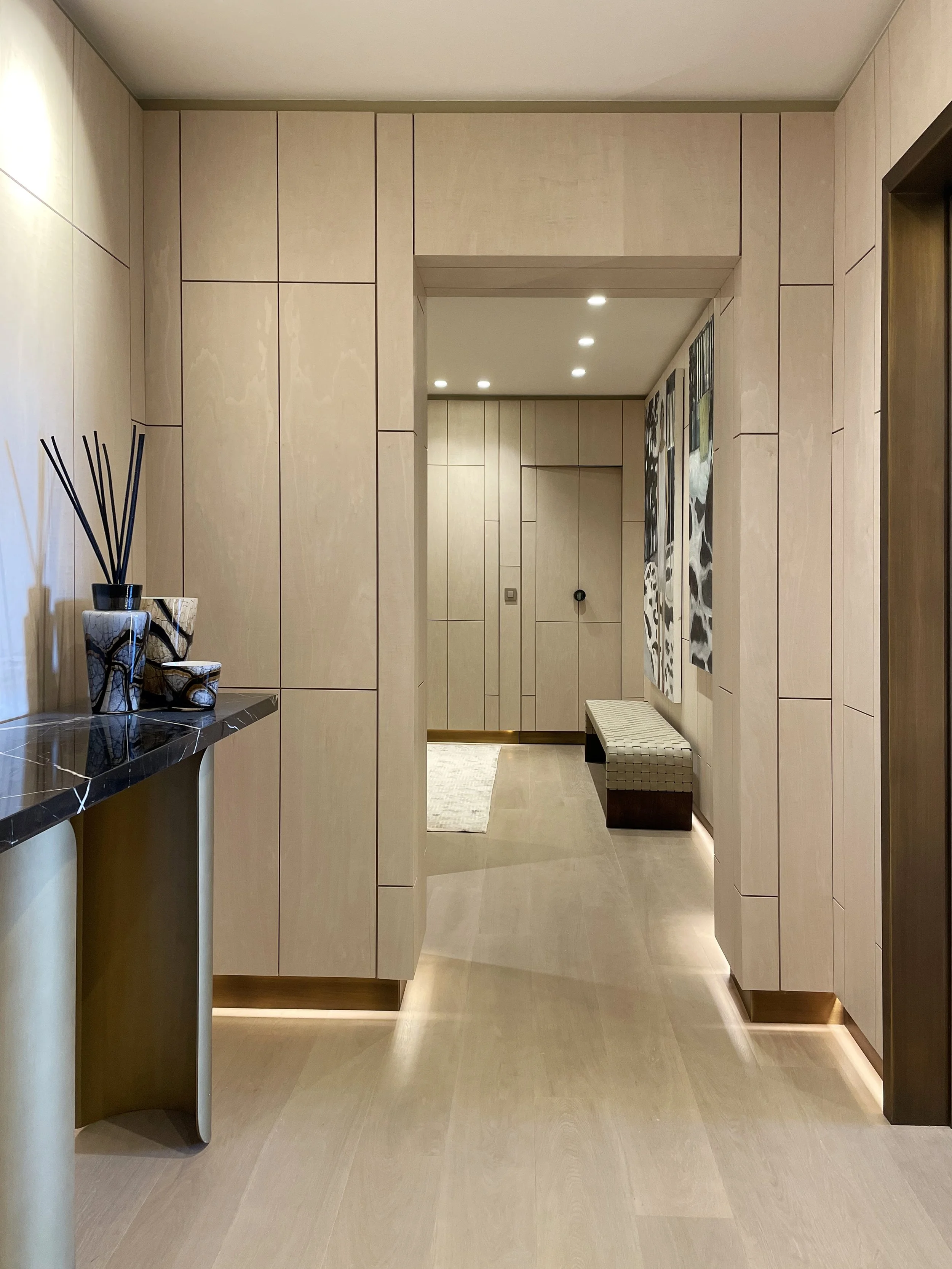 Un ingresso di una casa con pareti in legno chiaro, decorato con due vasi con bastoncini profumati a sinistra e un'area con appendiabiti e una panca con cuscino. Illuminazione a soffitto con faretti.
