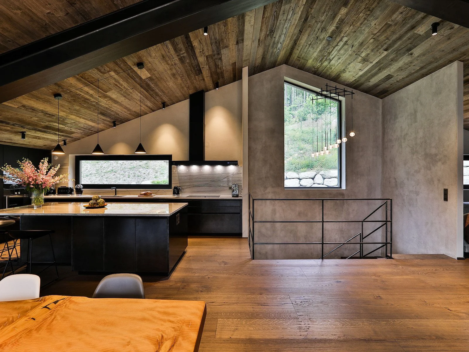 Interno moderno di una cucina con pareti in cemento, soffitto in legno, pannelli neri, piani di lavoro in marmo chiaro e un'isola centrale in legno con sedie nere. Decorazioni floreali e finestre che mostrano un ambiente naturale esterno.