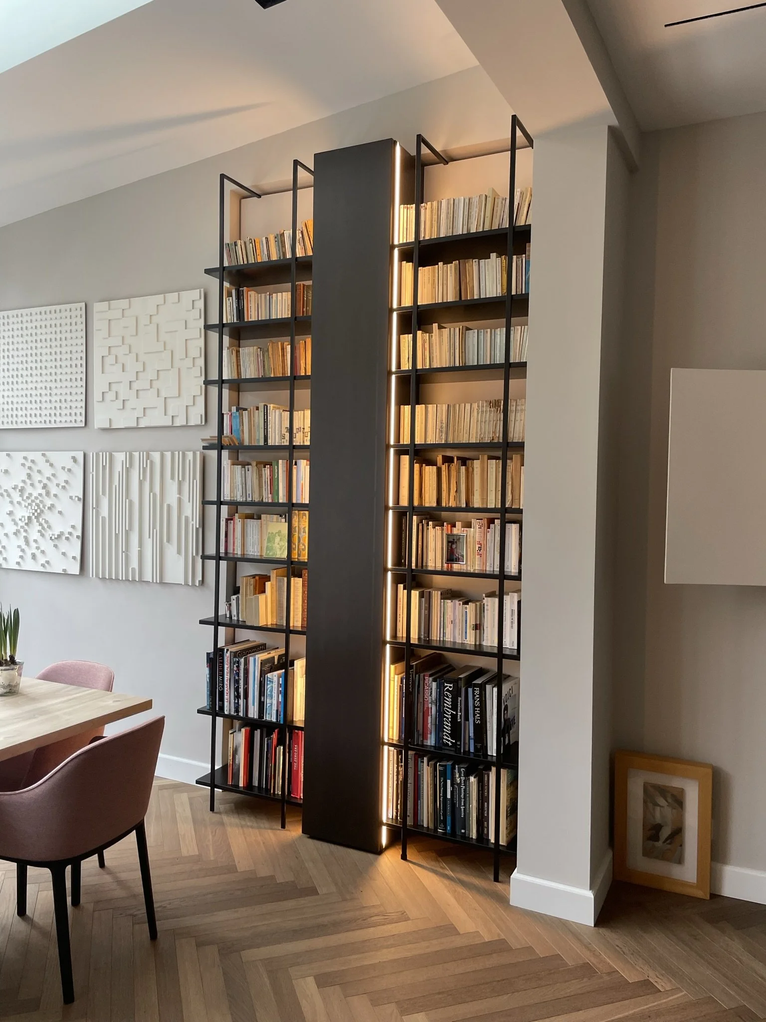 Scaffale in metallo nero con ripiani di libri colorati e bianchi, illuminato da luci LED, in un soggiorno con pareti chiare e opere d'arte decorativa. Parzialmente visibile un tavolo e sedie in primo piano, e un pavimento in legno a pavimento parquet.
