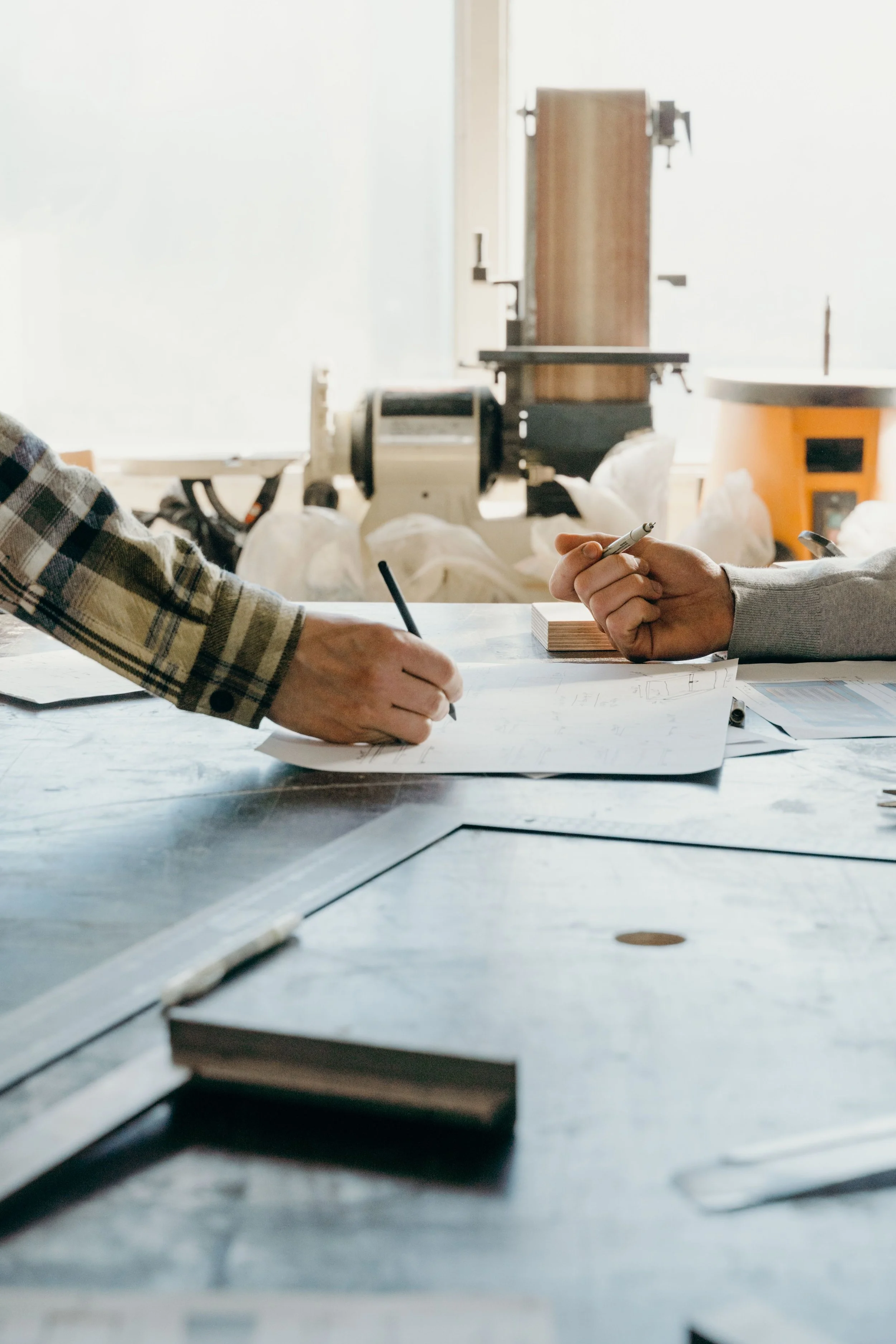 Due persone discutono o collaborano sui disegni e planimetrie su un tavolo da lavoro in un ambiente di progettazione o ufficio. Sul tavolo ci sono fogli, penne e strumenti di disegno.