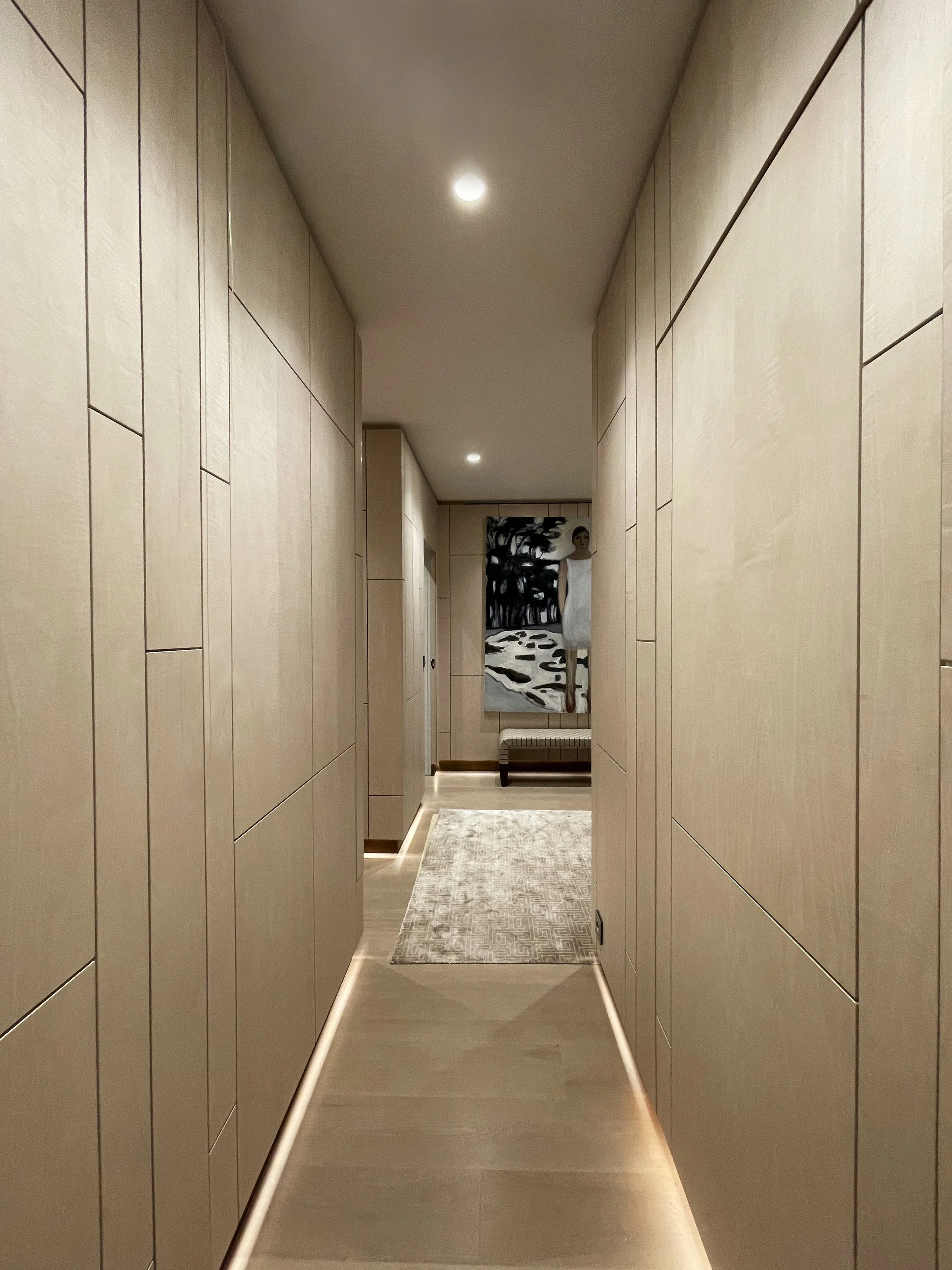 Un corridoio con pareti rivestite in legno chiaro, un tappeto bianco al centro, e un quadro d'arte moderna alla fine del corridoio, con un manichino vestito casualmente.