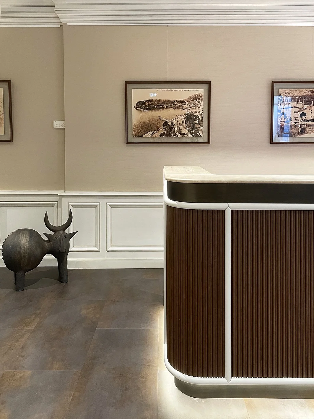 Dettaglio di un banco di reception in legno e piano di marmo chiaro. Si vedono quadri sulla parete  scultura di metallo appoggiata a terra a forma di bue.