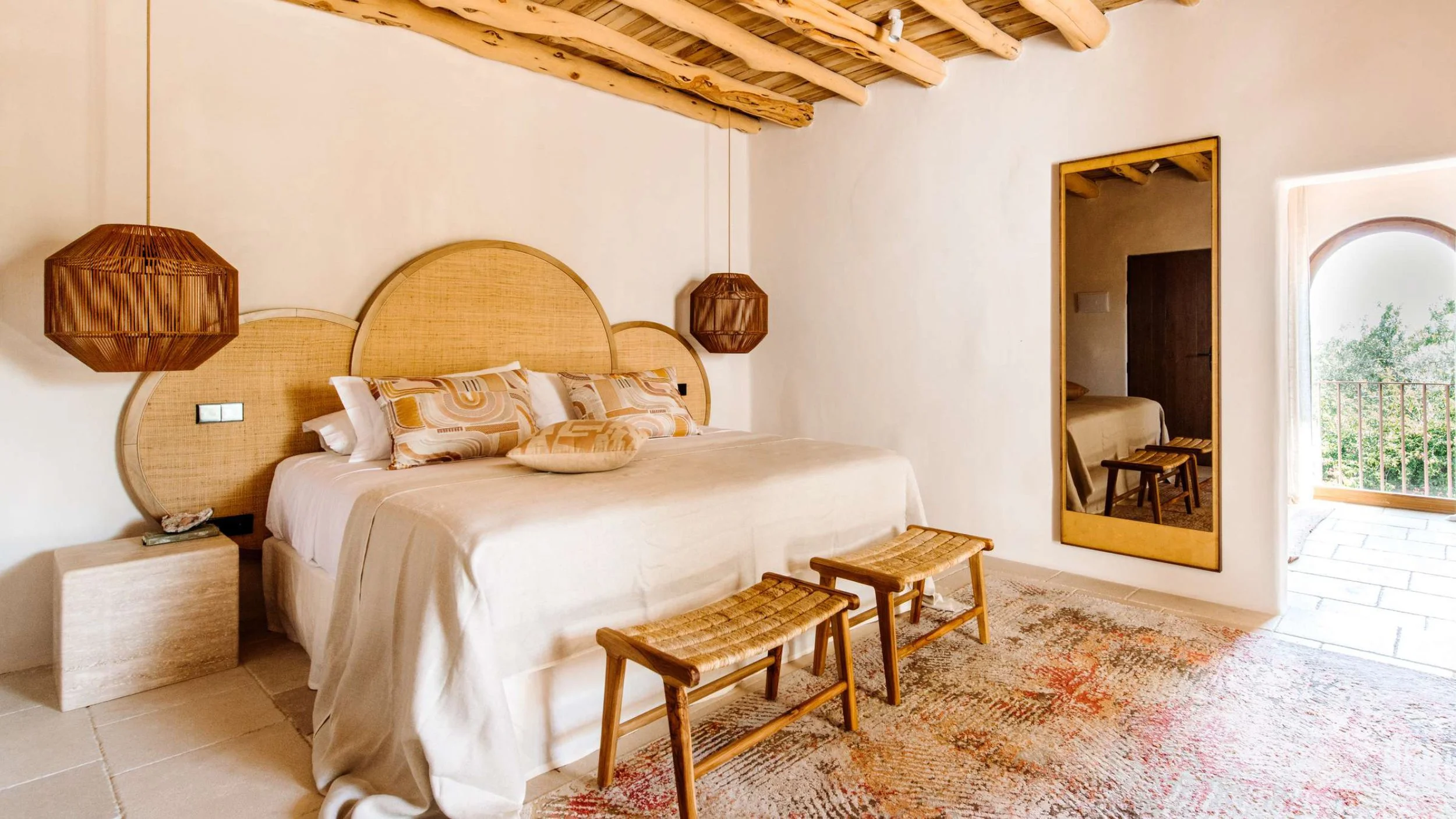 Camera da letto decorata in stile naturale con testa di letto in rattan, due lampade di vimini, parete bianca, grande specchio a muro, portafinestra con vista sul balcone e tappeto colorato.