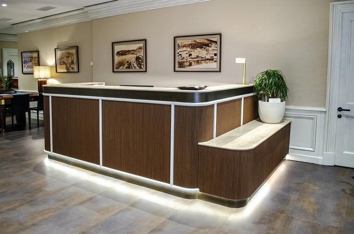 Banco della reception in legno e piano di marmo chiaro. Si vedono quadri sui muri e una pianta verde.