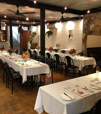 Elegant restaurant dining area with white tablecloths, floral centerpieces, and black chairs, featuring a stone fireplace and hanging plants.